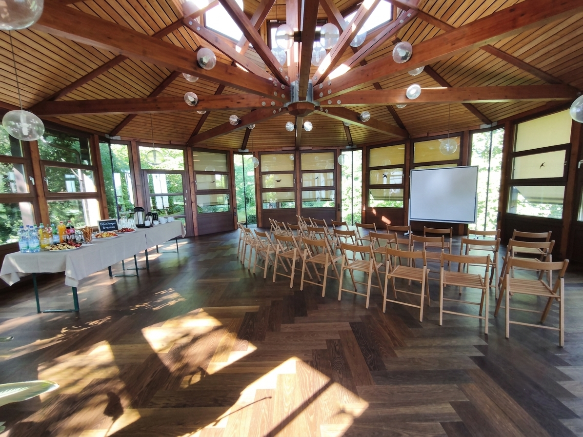 Ein lichtdurchfluteter, runder Veranstaltungsraum mit Holzdecke und großen Fenstern, Stühlen, einem Whiteboard und einem Buffet mit Getränken und Snacks.