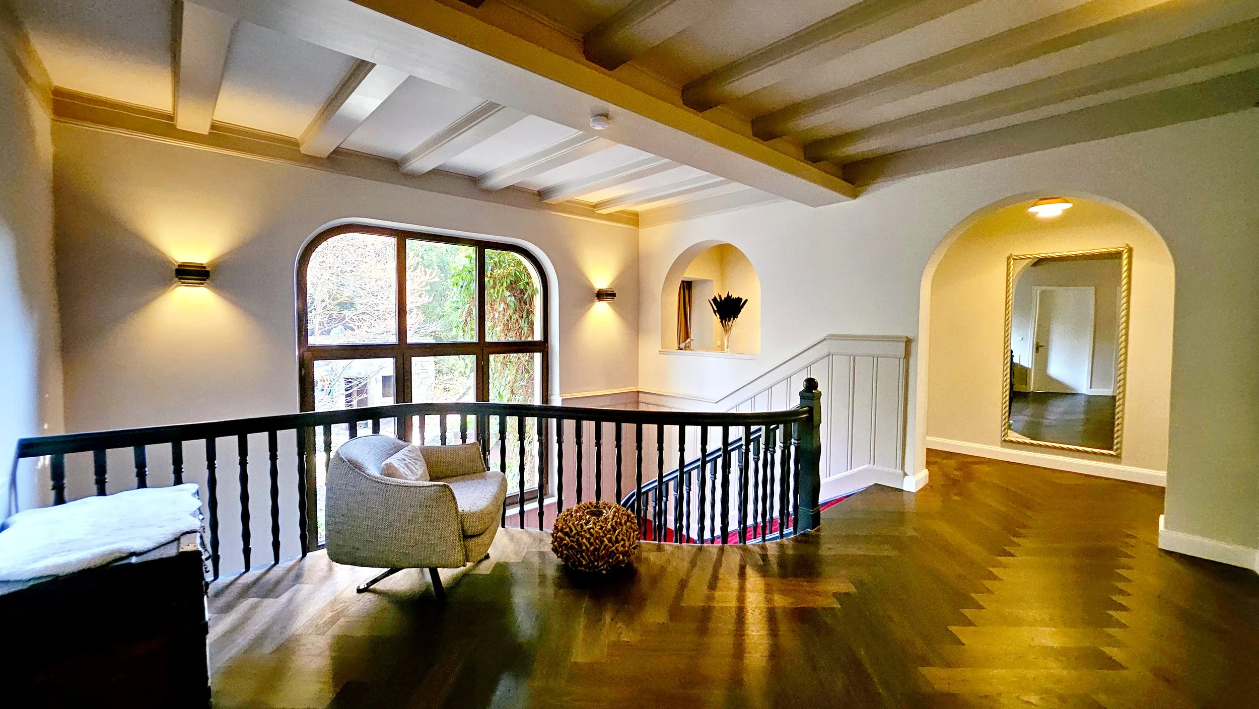 Ein heller, eleganter Wohnzimmerbereich mit Holzfußboden. Es gibt große Fenster, eine gepolsterte Sitzgelegenheit, einen Sessel, einen kleinen Sitzhocker, einen Spiegel mit goldfarbenem Rahmen, Wandlampen und eine dekorative Vase. Der Raum ist lichtd