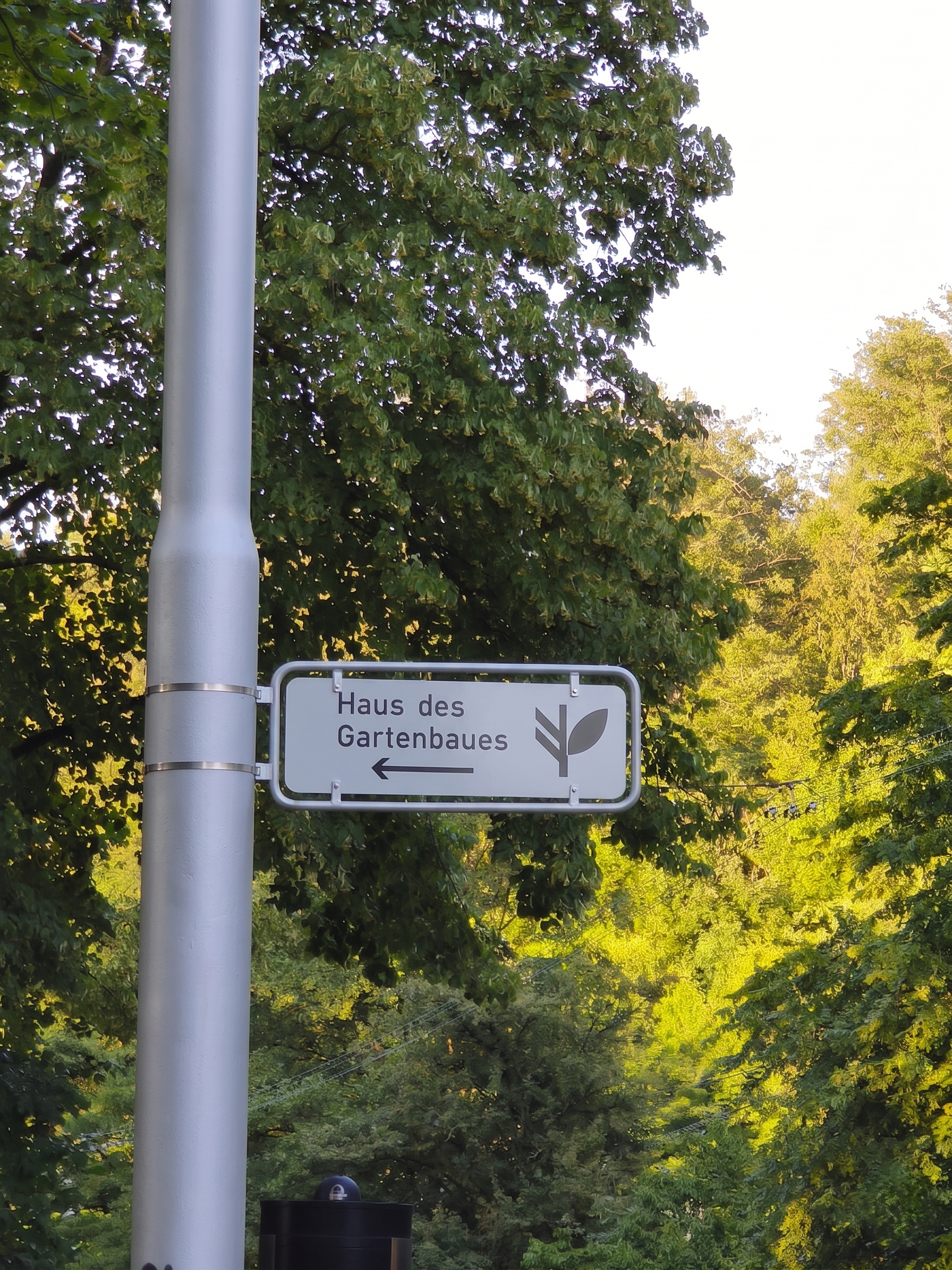 Ein Straßenschild mit der Aufschrift 'Haus des Gartenbaus', einem Pfeil nach links, und einem Blatt-Logo, umgeben von grünen Bäumen im Freien.