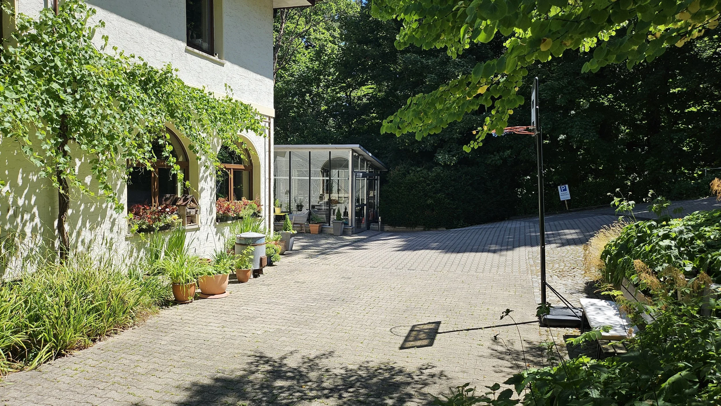 Ein gepflegter Gartenbereich mit Blumen und Pflanzen vor einem weißen Haus, eine Sitzecke mit Glasüberdachung und ein Parkplatz mit einem Parkplatz-Schild.