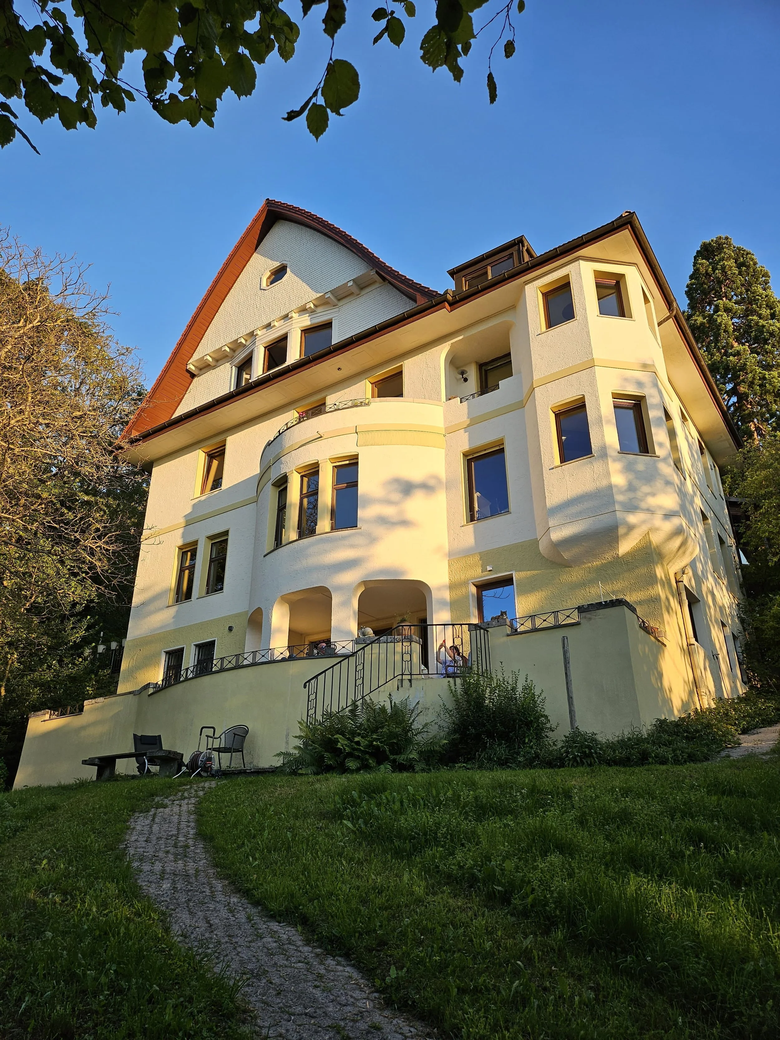 Mehrstöckiges Haus mit weißen Wänden, mehreren Fenstern und Balkon, umgeben von Bäumen und grünem Gras, bei Sonnenuntergang, blauer Himmel