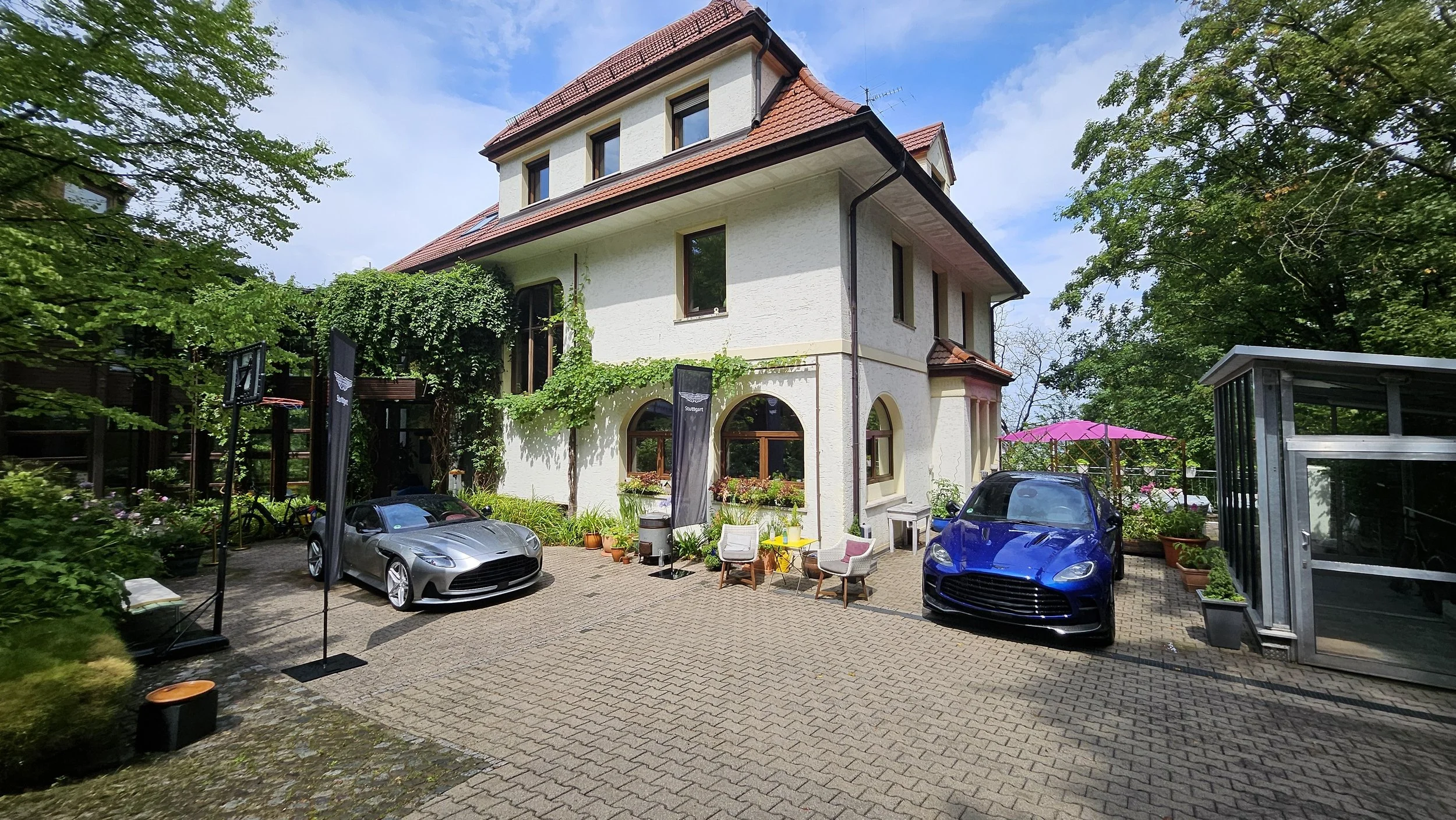 Mehrstöckiges weißes Haus mit rotem Satteldach, umgeben von Bäumen und Pflanzen, im Vordergrund ein gepflasterter Hof mit zwei Autos, Sitzmöbeln, Sonnenschirm und verschiedenen Dekorationen.