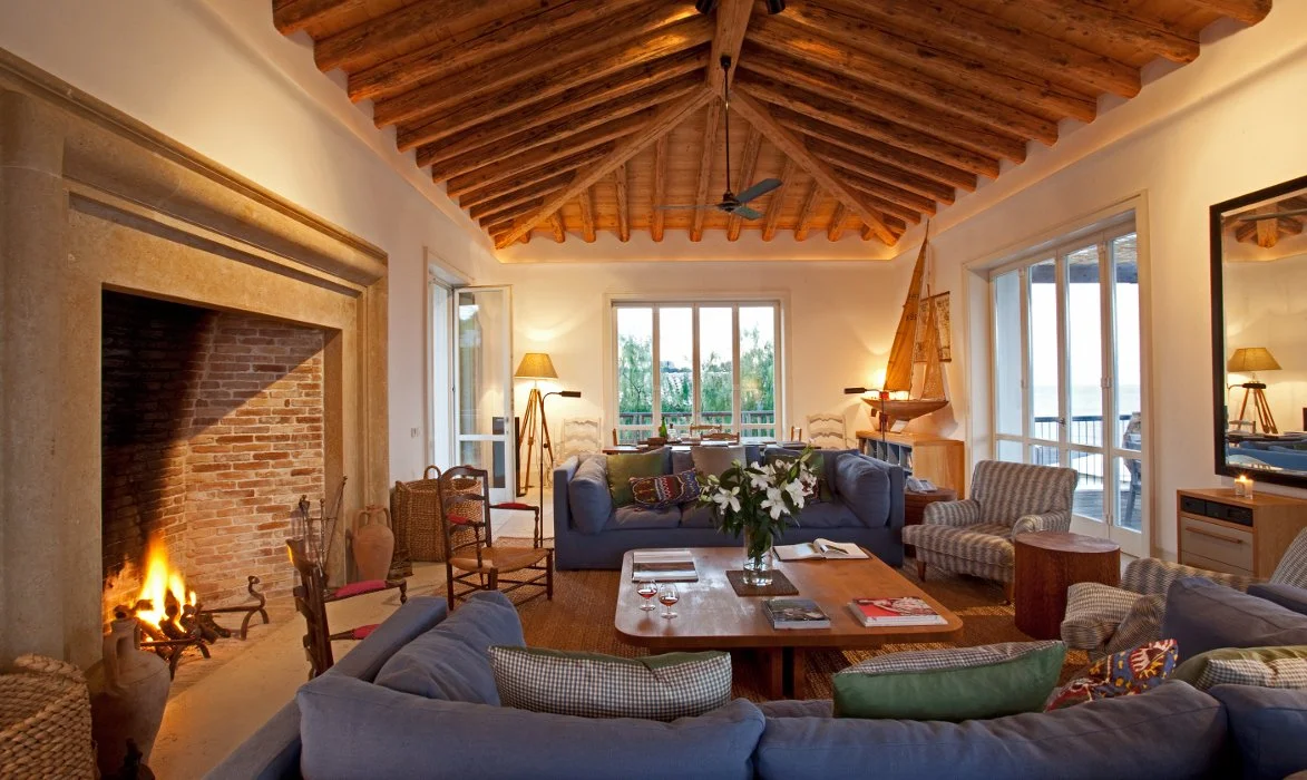 Living room with a wood-beamed ceiling, a fireplace with a fire, large windows, and various sofas and chairs around a wooden coffee table with flowers.
