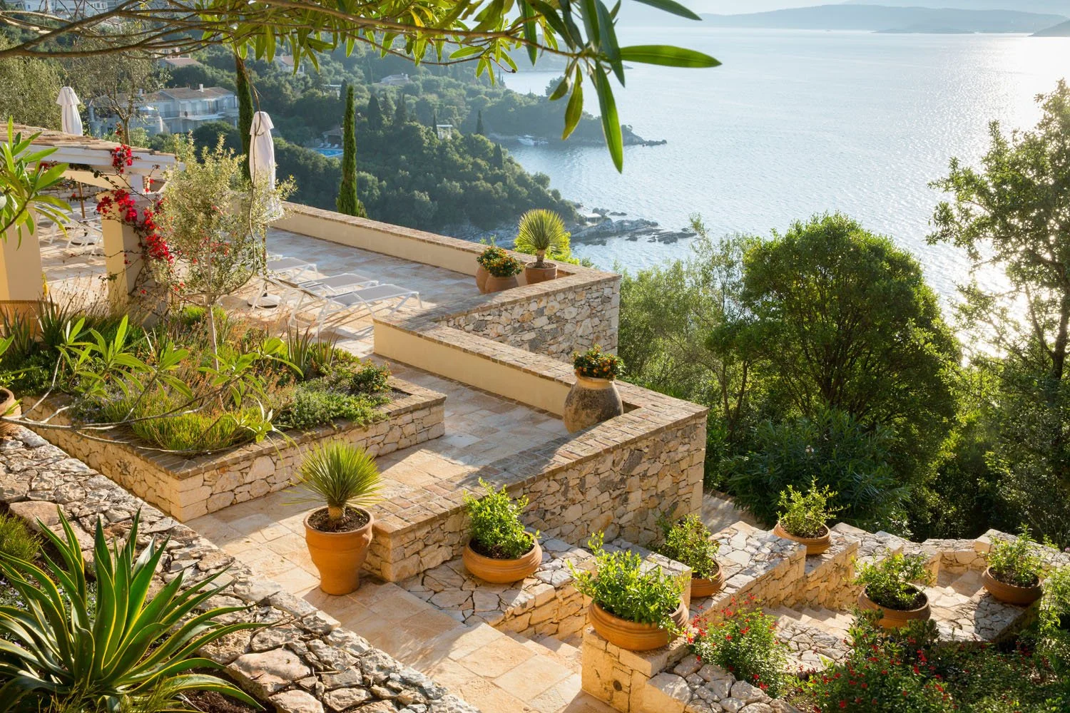 A terraced garden with stone walls and potted plants overlooking a body of water and a lush landscape.