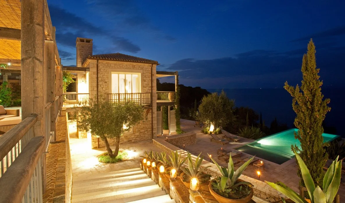 A luxurious house with outdoor lighting, a swimming pool, and a view of the ocean at dusk.
