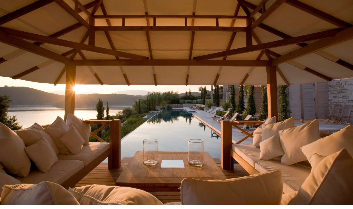 A luxurious outdoor poolside lounge area with cushioned seating under a large wooden gazebo, overlooking a river sunset with hills in the background.
