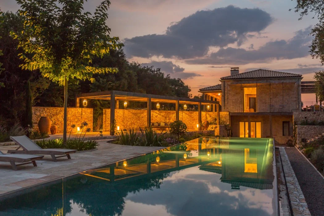 A luxurious outdoor swimming pool area at dusk with lounge chairs, lanterns, and a modern house in the background.