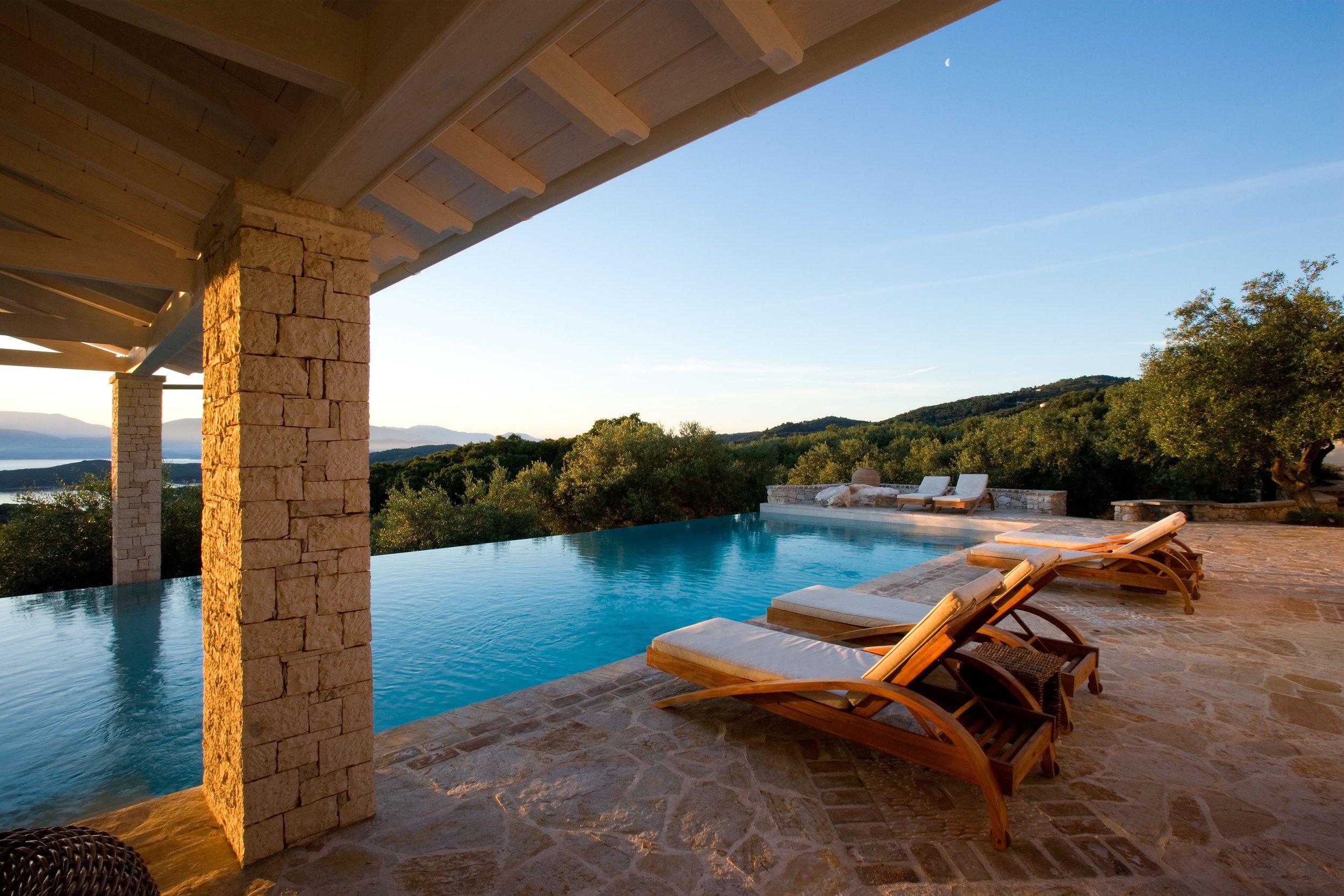 A luxurious outdoor infinity pool with wooden lounge chairs on a stone deck, overlooking lush green hills and a body of water under a clear blue sky with a visible moon.