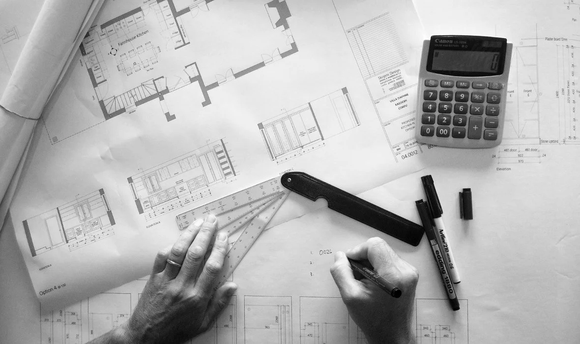 Architectural plans and drawings on a table with tools, pens, a calculator, and a person's hand measuring with a ruler.