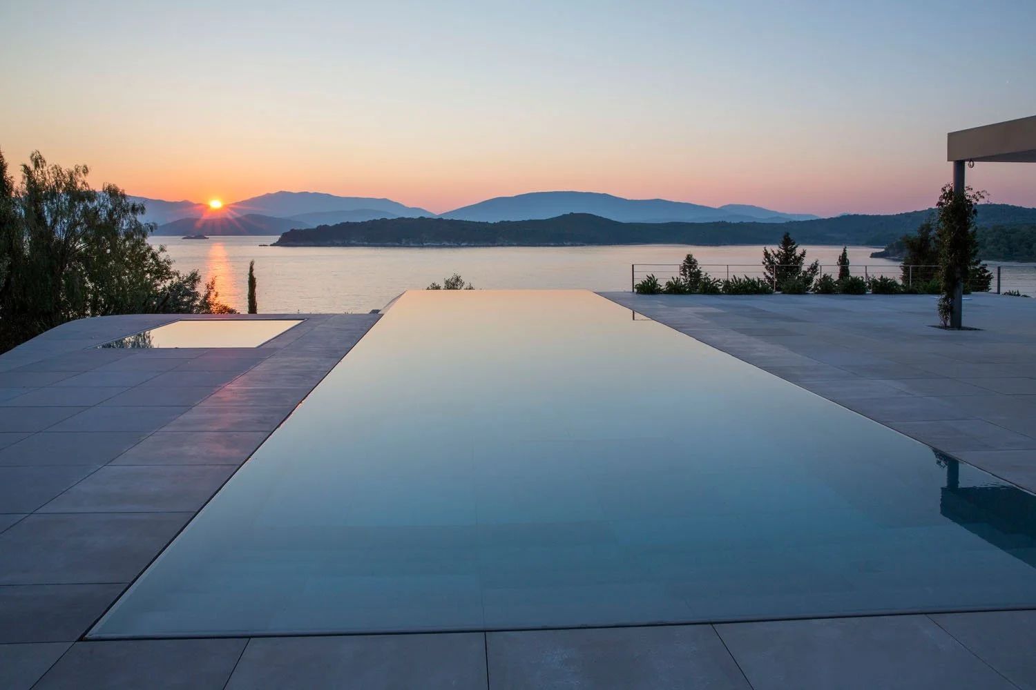 A modern infinity pool overlooking a lake with mountains in the distance during sunset.