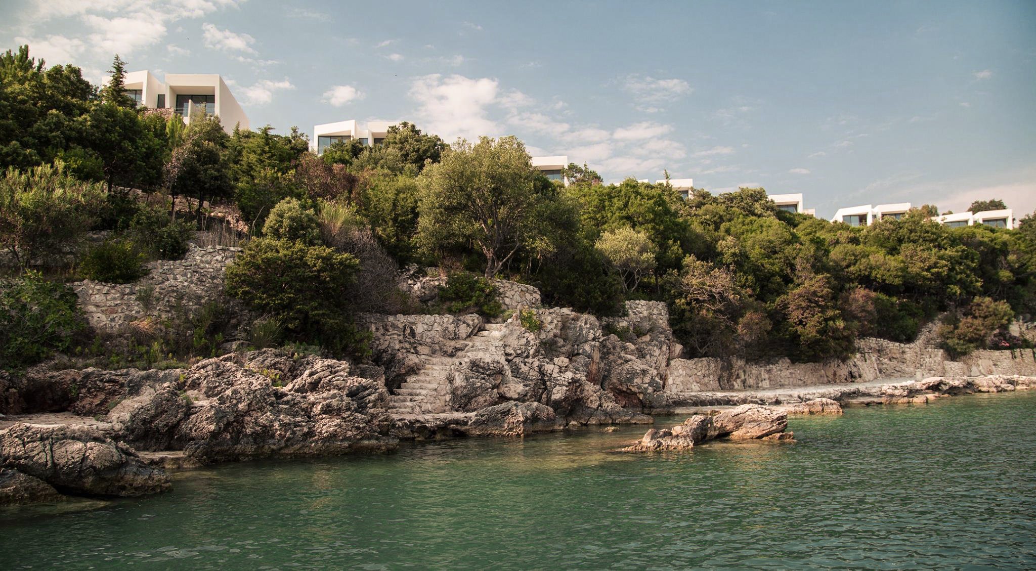 Modern white houses situated on a hillside lush with green trees, overlooking rocky shoreline and calm water with a cloudy sky overhead.