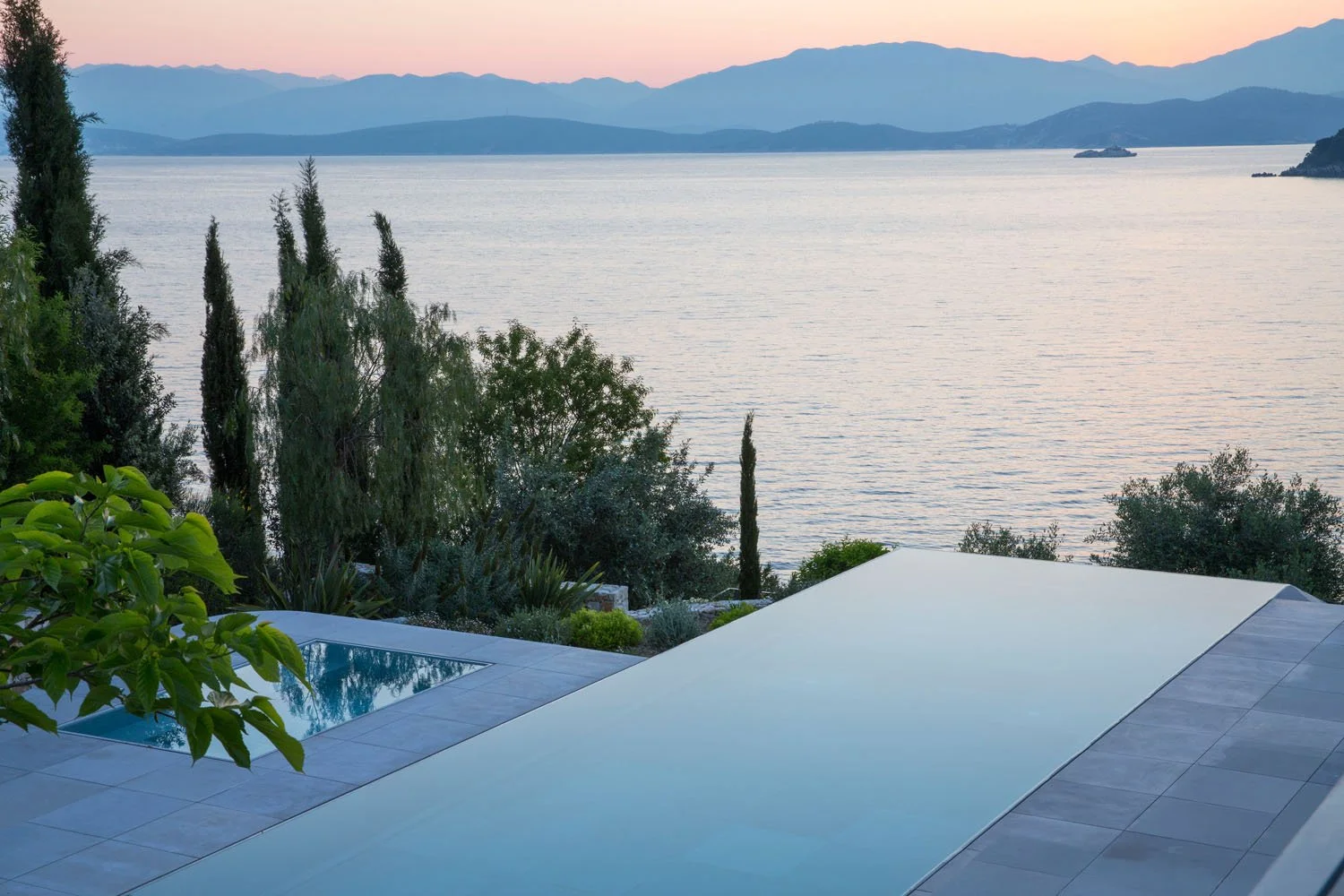 View of a serene lake with distant mountains at sunset, outdoor pool and lush greenery in the foreground.
