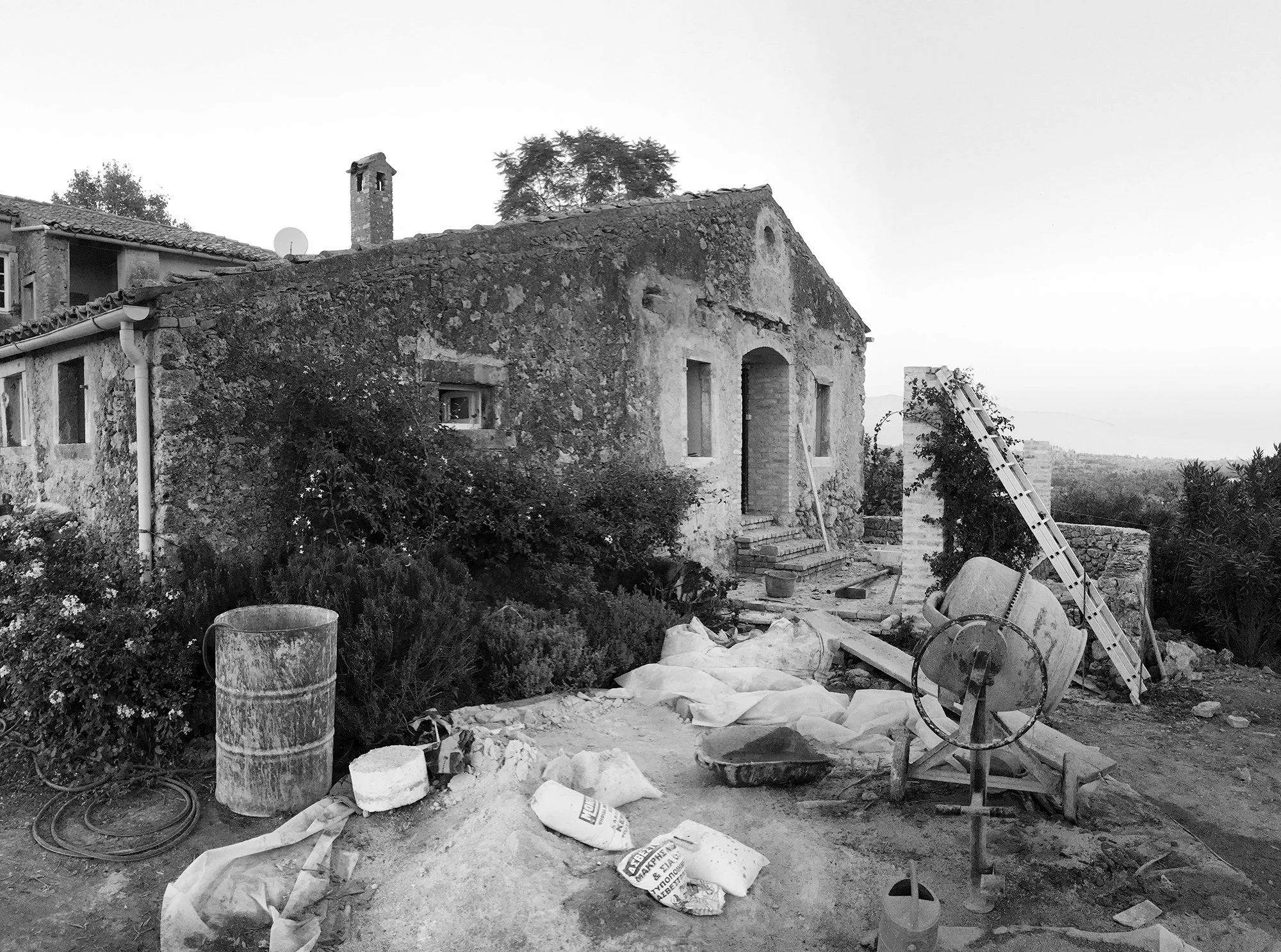 A black-and-white photo of a rustic house with renovation work in progress. The house has stone walls, arched doorway, and small windows. Construction tools and materials, including a ladder, barrels, and bags, are scattered in the yard, indicating ongoing building or restoration work.