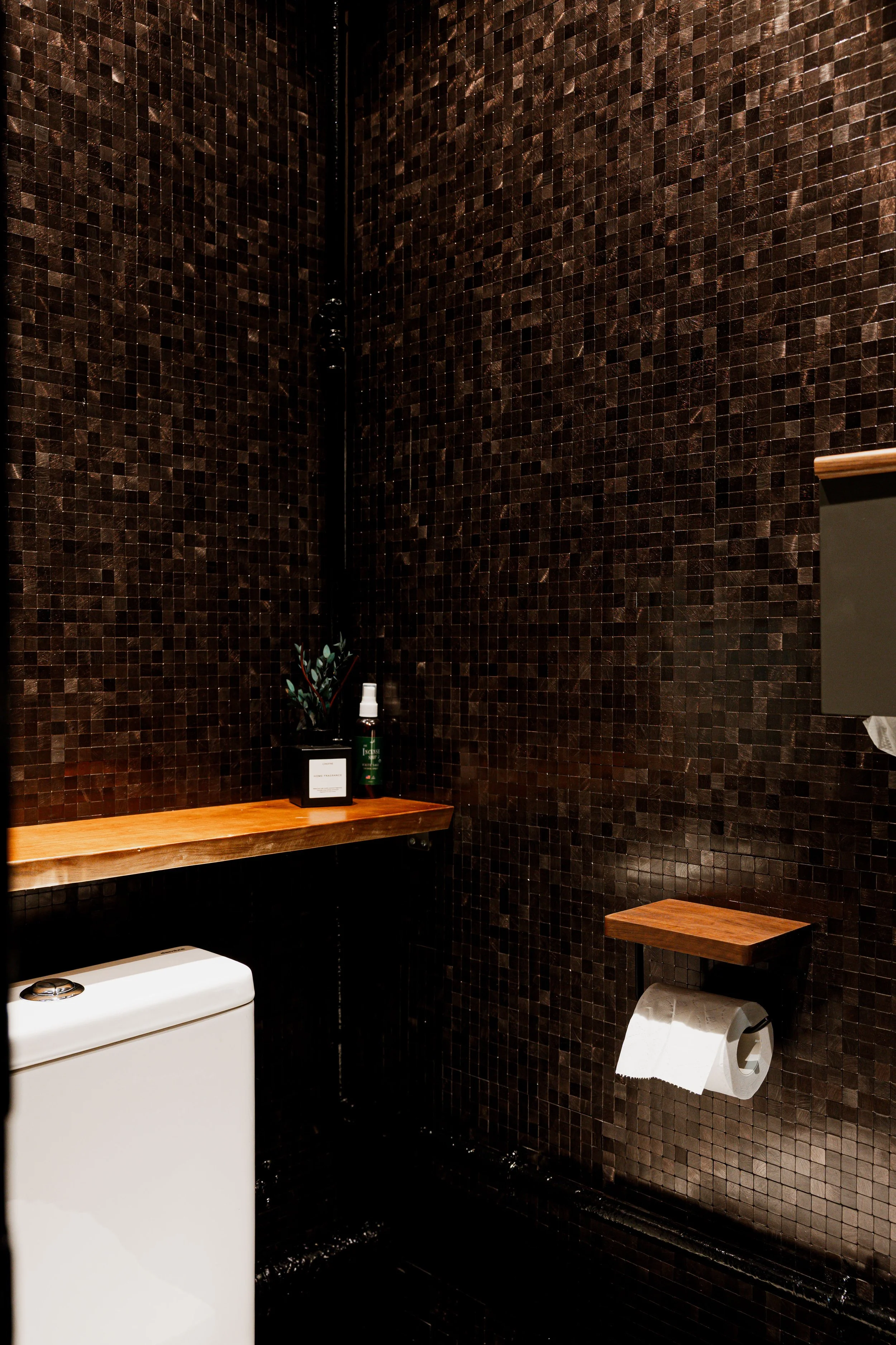 A modern bathroom corner with dark brown mosaic tile walls, a wooden shelf, a small plant, a spray bottle, a box, a white toilet, and a toilet paper roll holder.