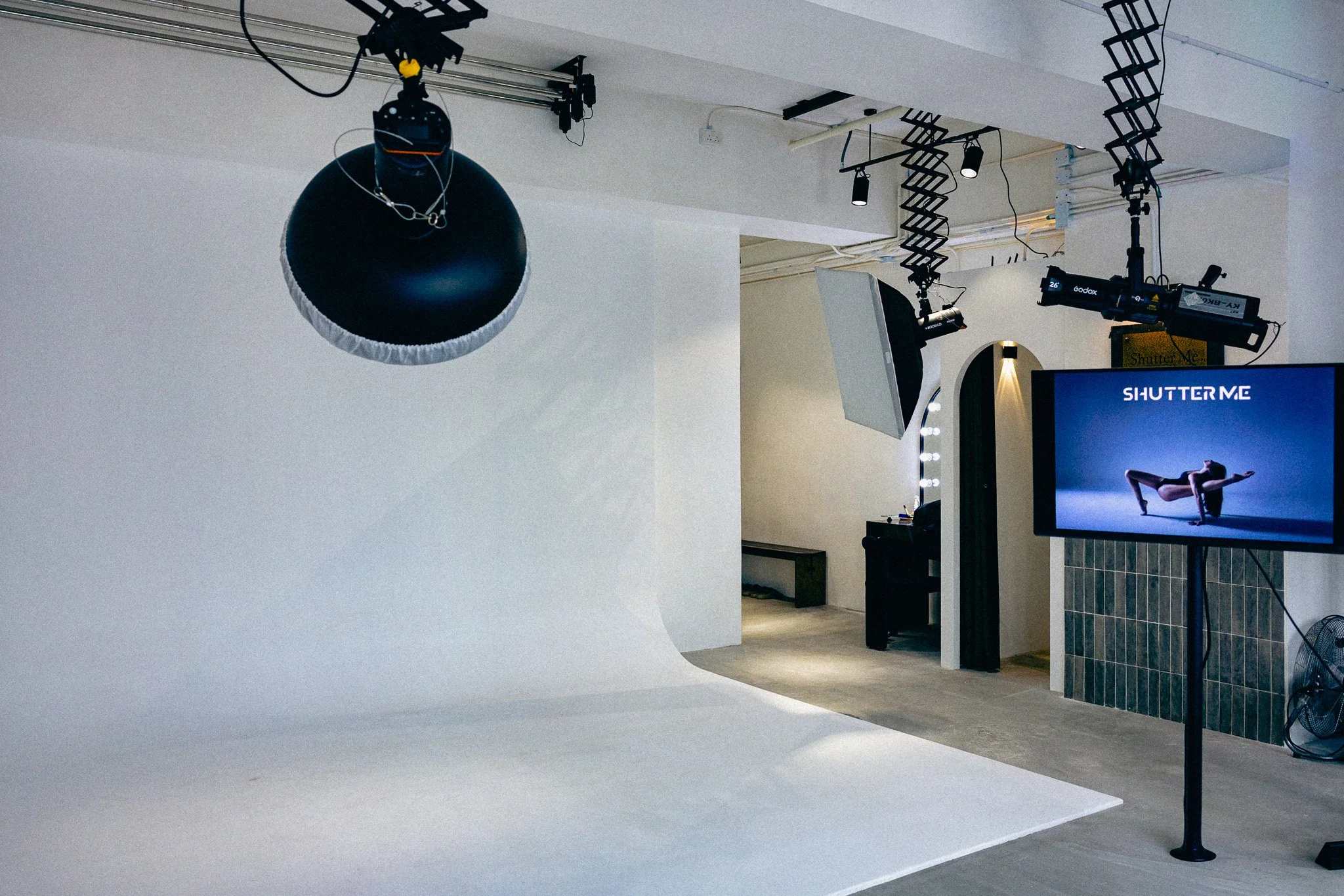 Photography studio with a white seamless background, professional lighting equipment including a large umbrella light, and a digital display screen showing an image of a woman doing a yoga pose.