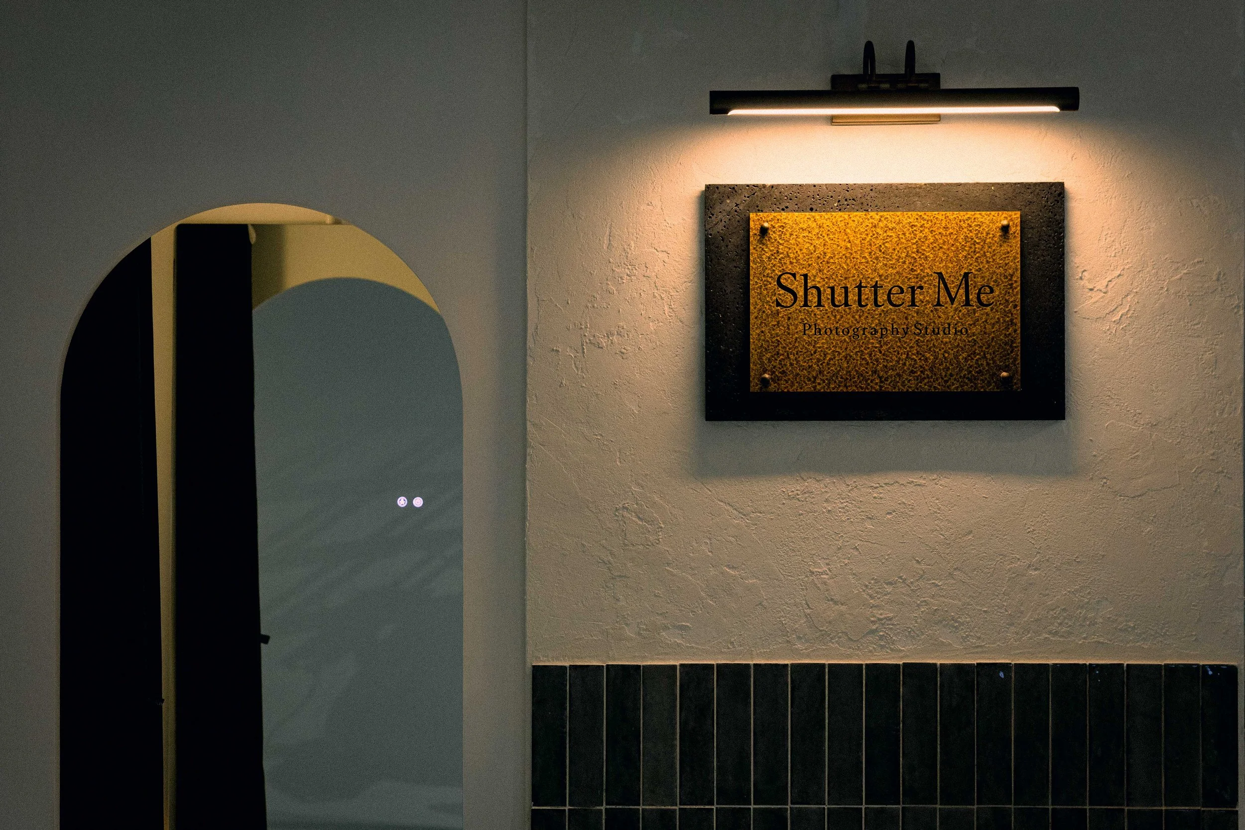 A wall-mounted sign reading 'Shutter Me Photography Studio' illuminated by a horizontal light fixture. Part of a mirror is visible on the left side, and part of a tiled wall is at the bottom of the image.