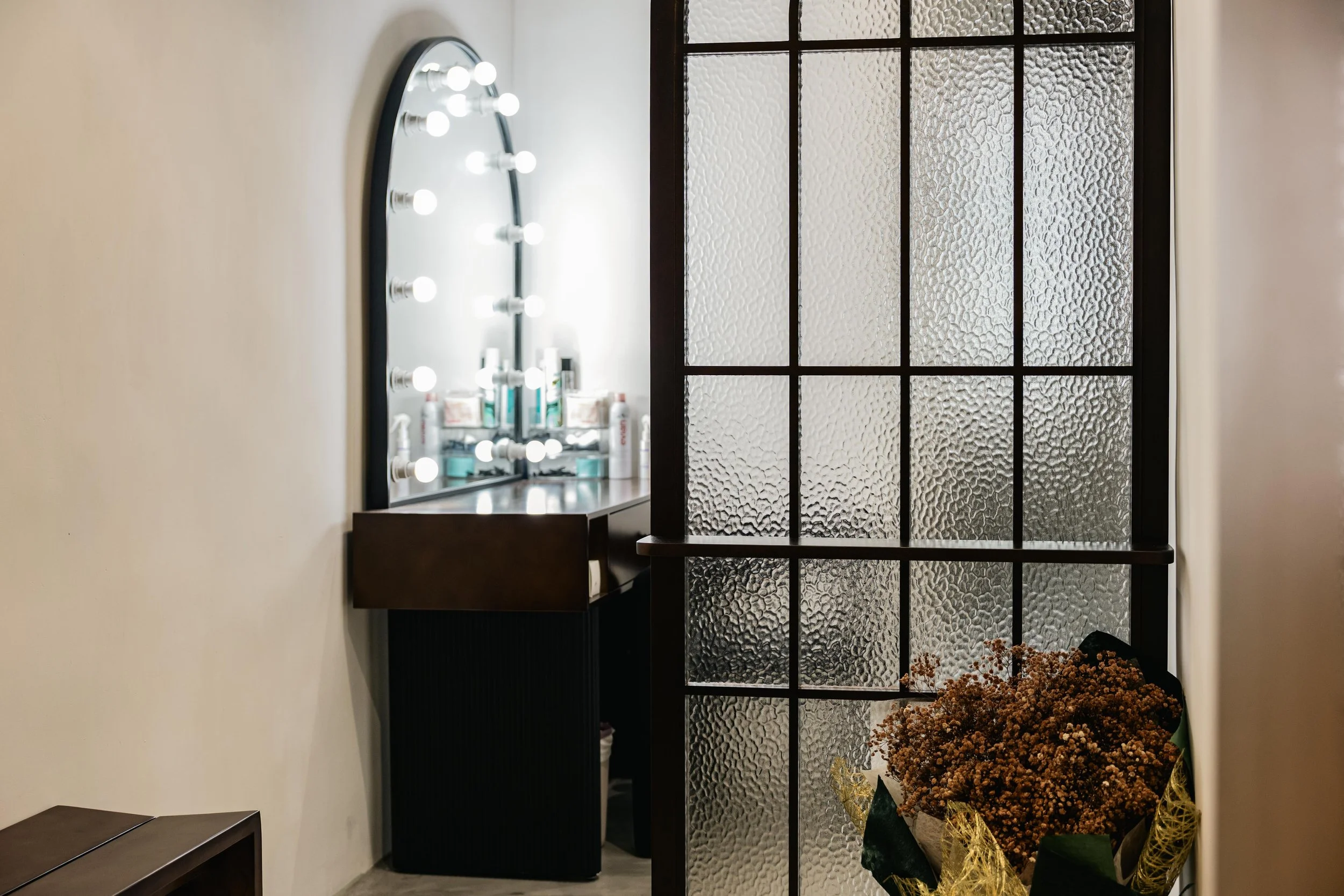 A room with a circular mirror with lights on the wall, a textured glass window, a black side table, and a large pot of dried flowers.