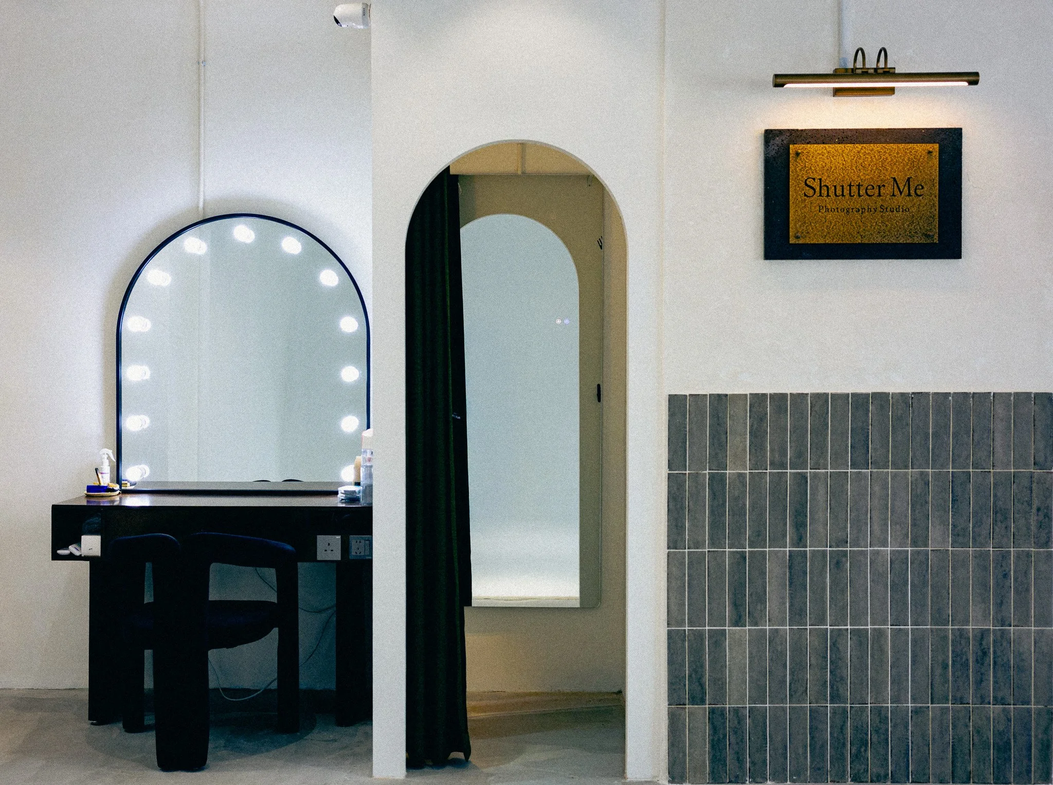 Interior view of a photography studio with a large illuminated mirror, a black table, chairs, and a sign that reads 'Shutter Me Photography Studio' on the wall. There is a doorway with a mirror and a curtain.