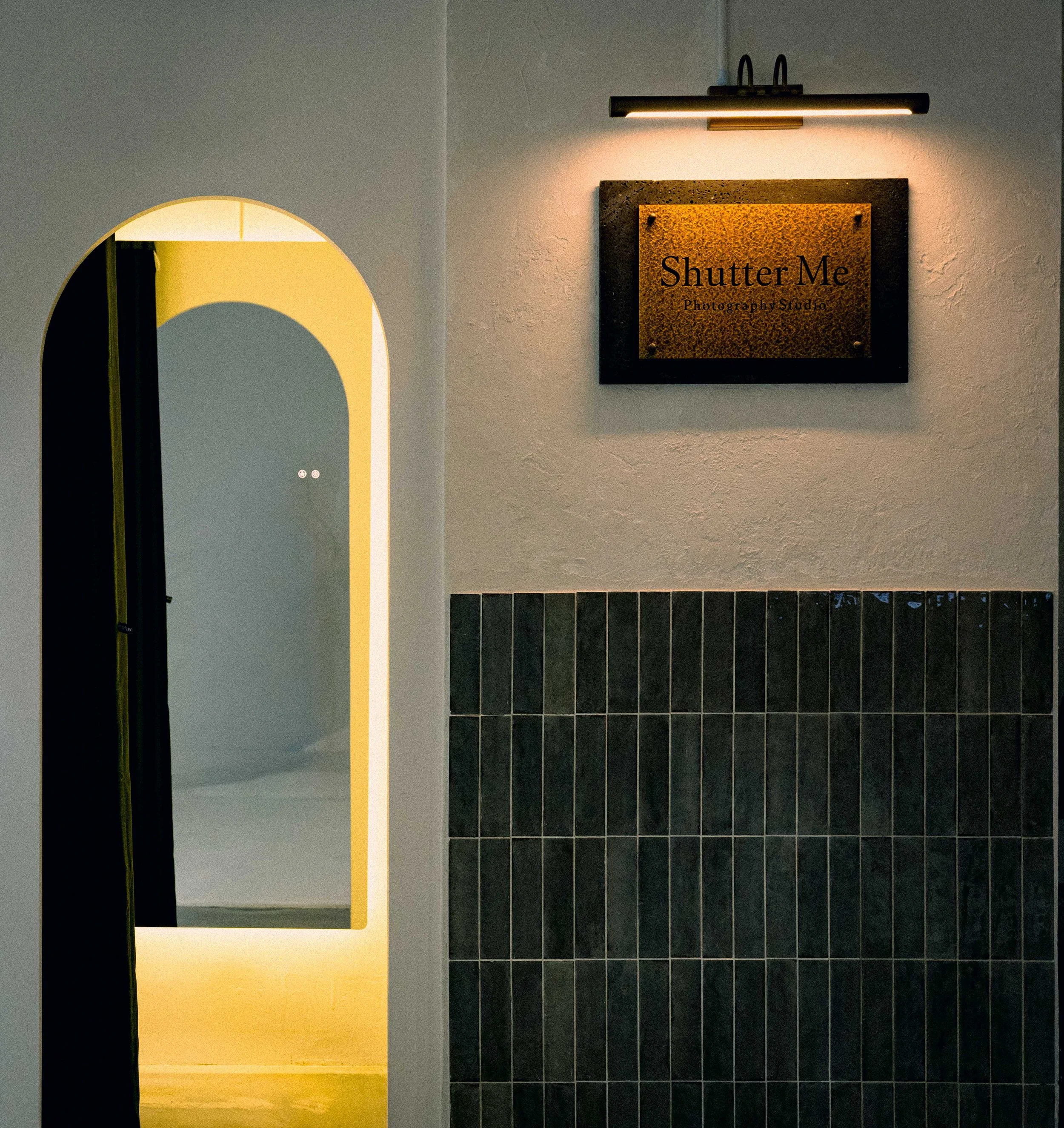 Sign for Shutter Me Photography Studio hanging on a white wall, illuminated by a black wall-mounted light fixture, next to a mirror with a rounded top edge and black trim in a room with dark tiled lower wall.