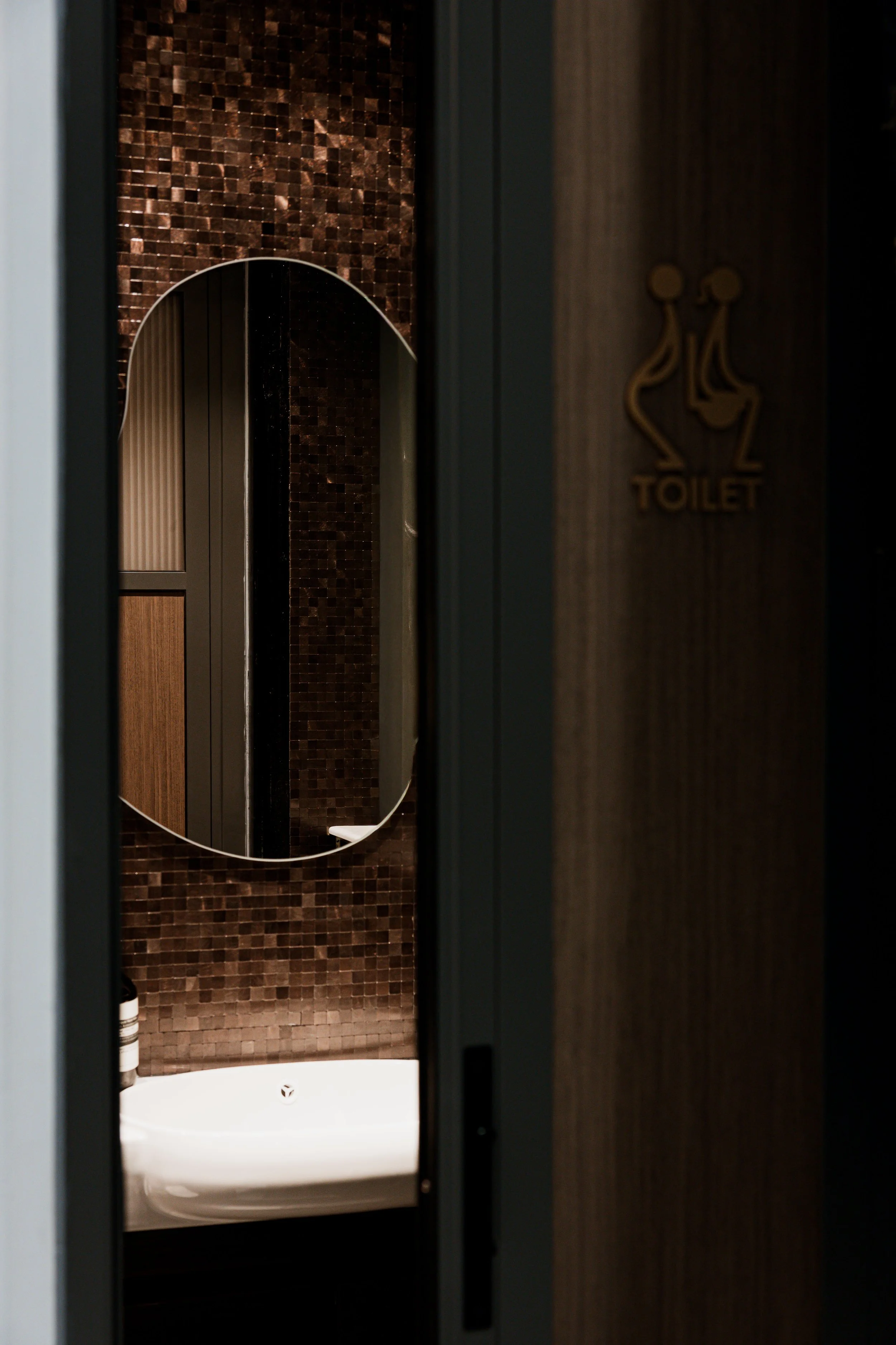 A view into a bathroom with a mirror, brown mosaic tiles, and a sign with a stylized elegant image of two women and the word 'TOILET' on the wall.