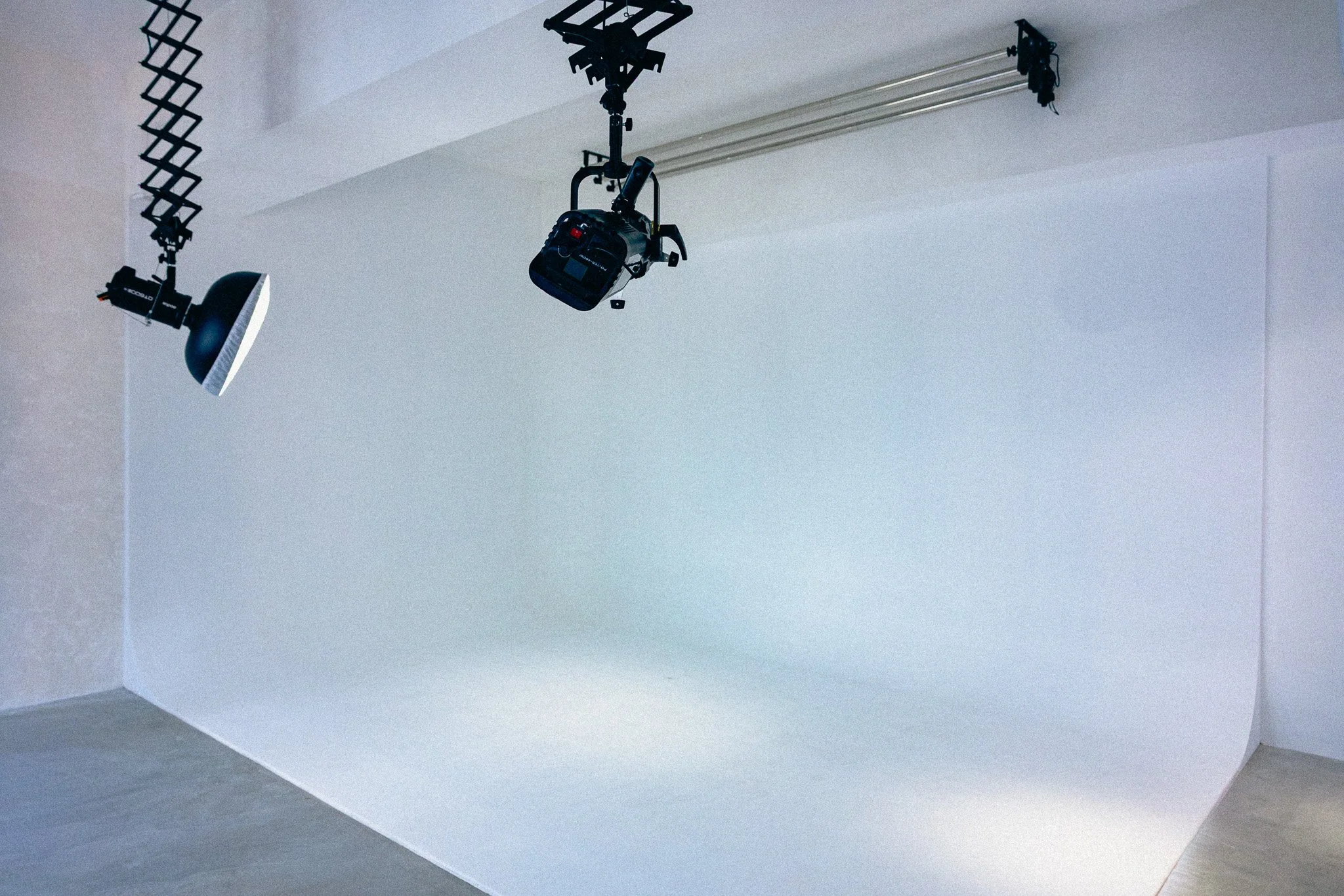 Photo of a white corner of a room with professional film or photography lighting equipment hanging from the ceiling, including a large softbox light on a stand and a grid-like light modifier.