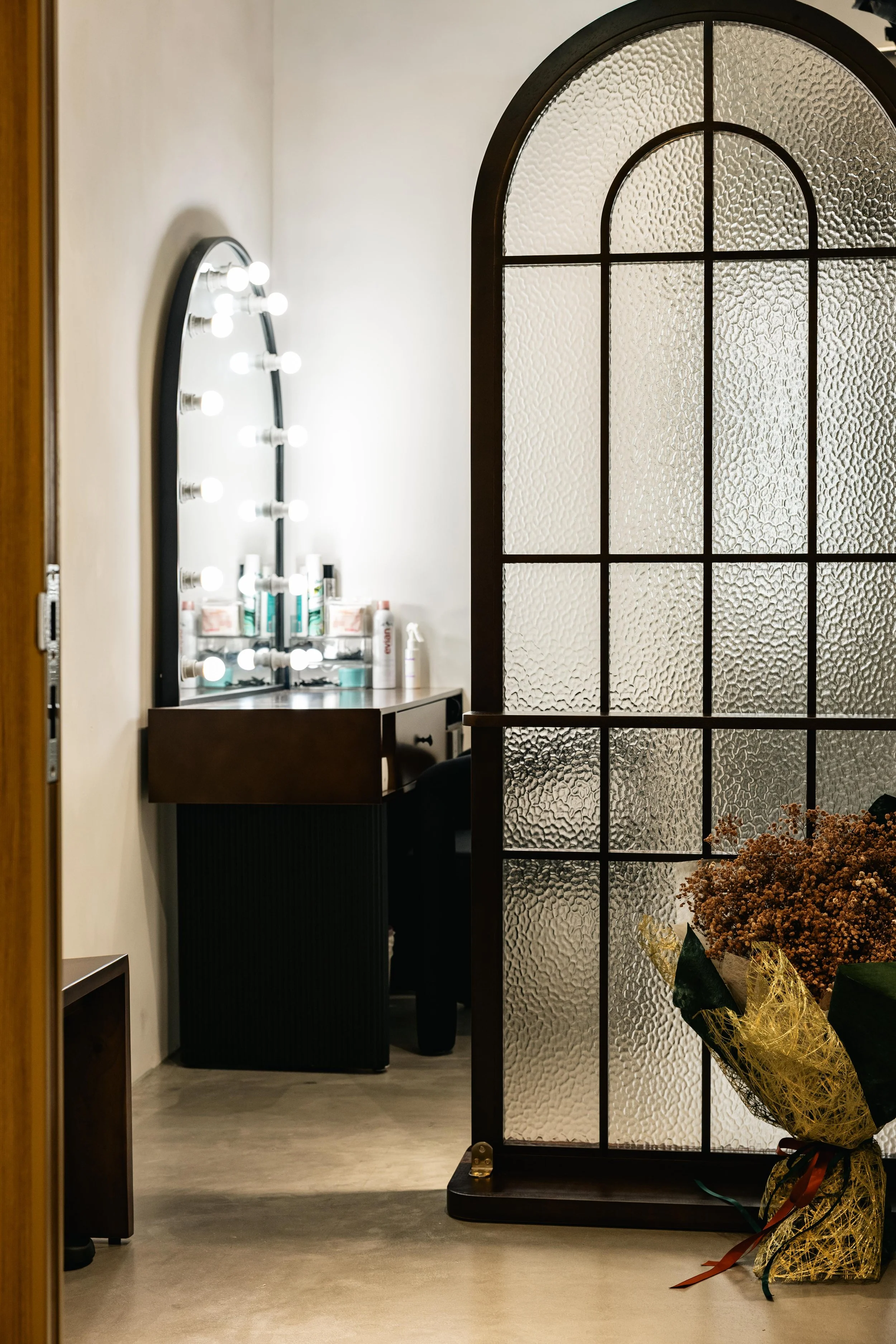 Interior of a room with a white wall mirror surrounded by light bulbs, a wooden drawer, textured glass divider, and a bouquet of brown flowers wrapped in gold and green wrapping paper.