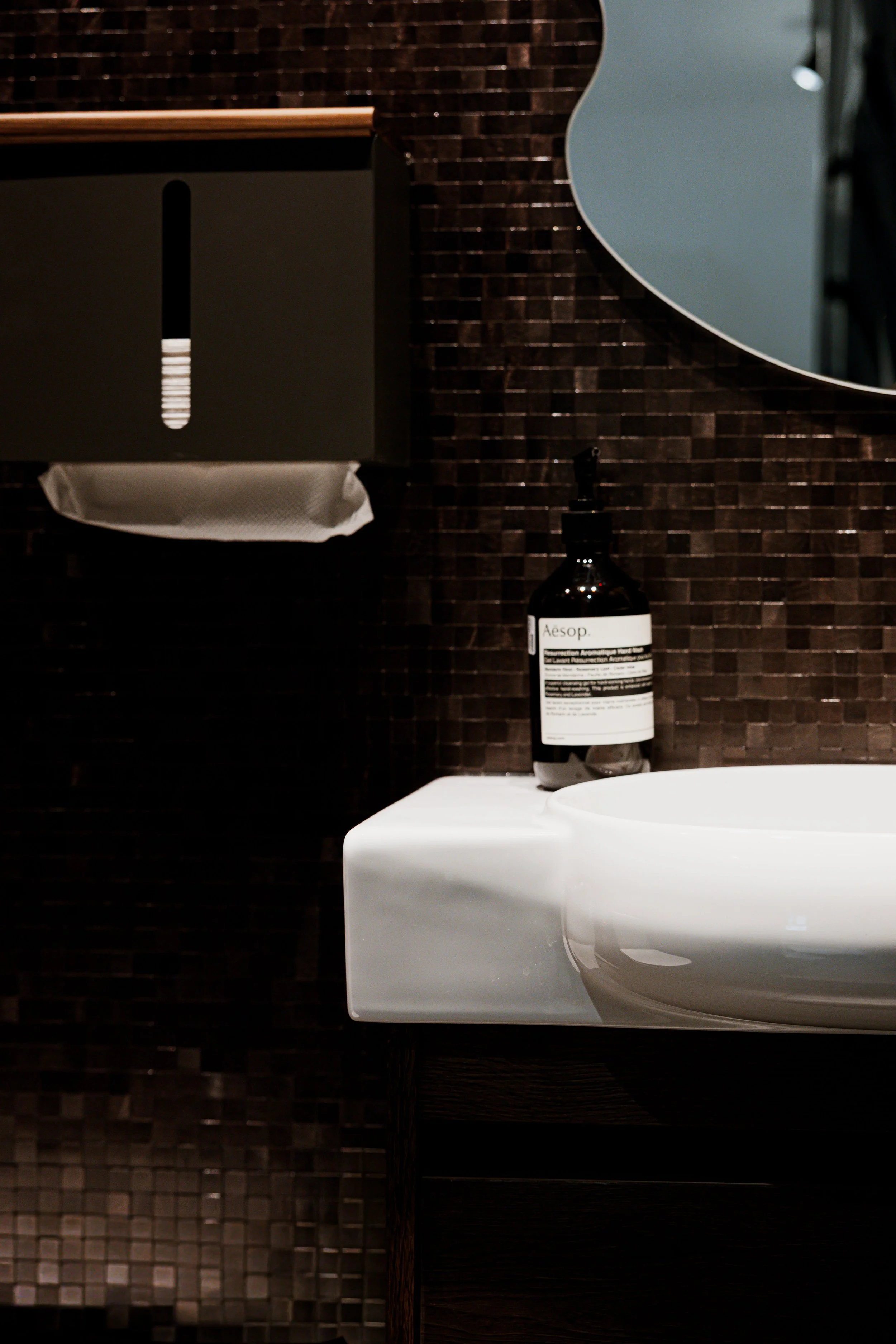 Close-up of a modern bathroom counter with a white sink, a black soap dispenser labeled 'Aesop,' and a dark tiled wall with a mirror reflecting part of the room.