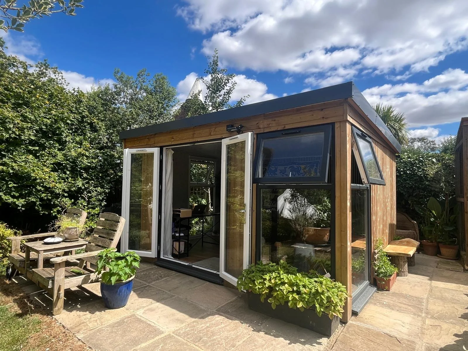 A small wooden garden shed with large windows and open double doors, surrounded by potted plants and outdoor furniture, under a partly cloudy sky.