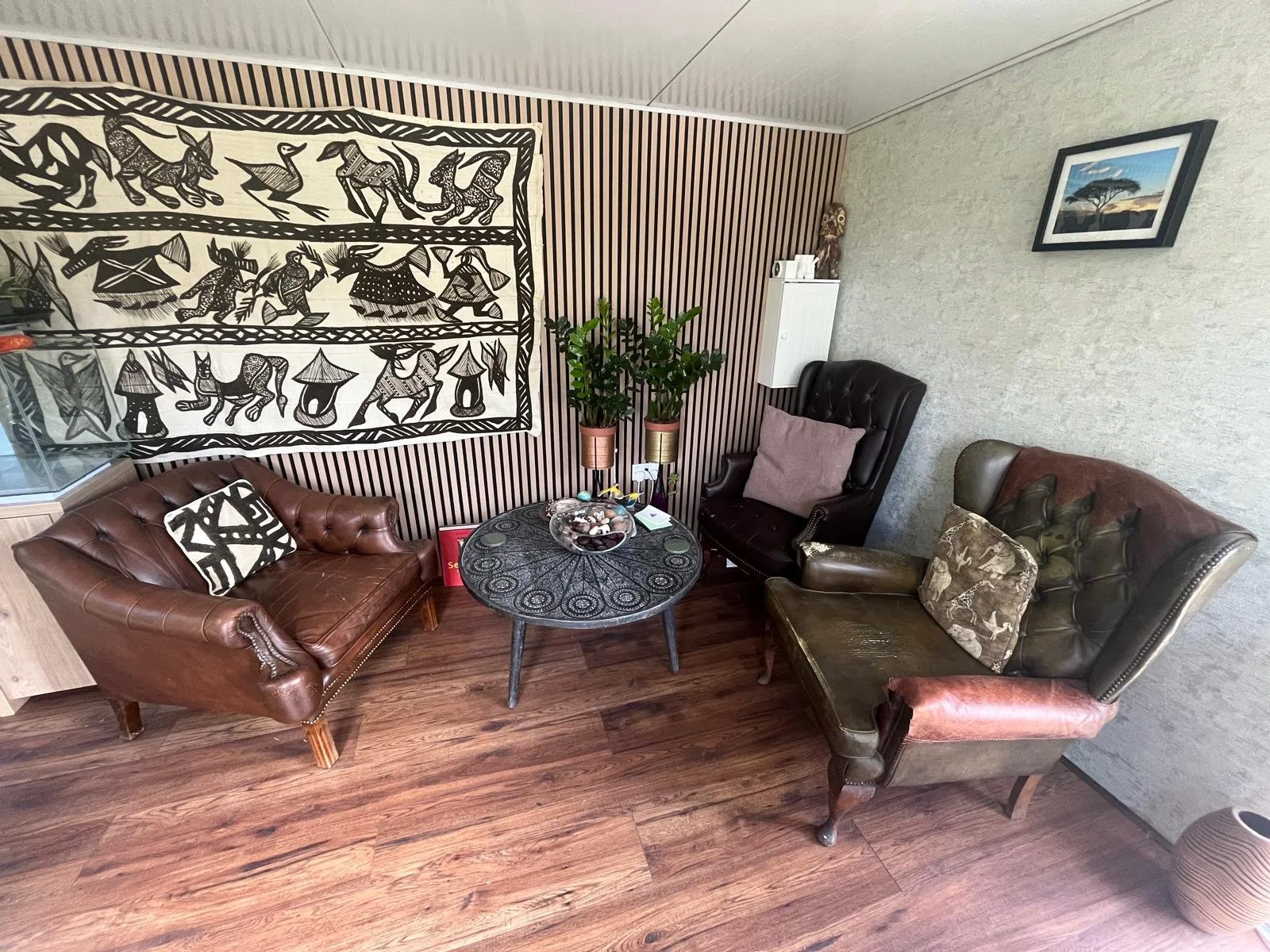 A cozy living room with a brown leather sofa, a black high-back armchair, and a dark green armchair, all with decorative pillows. There is a round black coffee table in the center with shells and decorative items. Two potted plants sit near a wall with a striped pattern. The wall on the right has a framed landscape picture, and a white cabinet with a carved wooden sculpture on top. The floor is wood, and there is a large black-and-white tribal art piece on the back wall.
