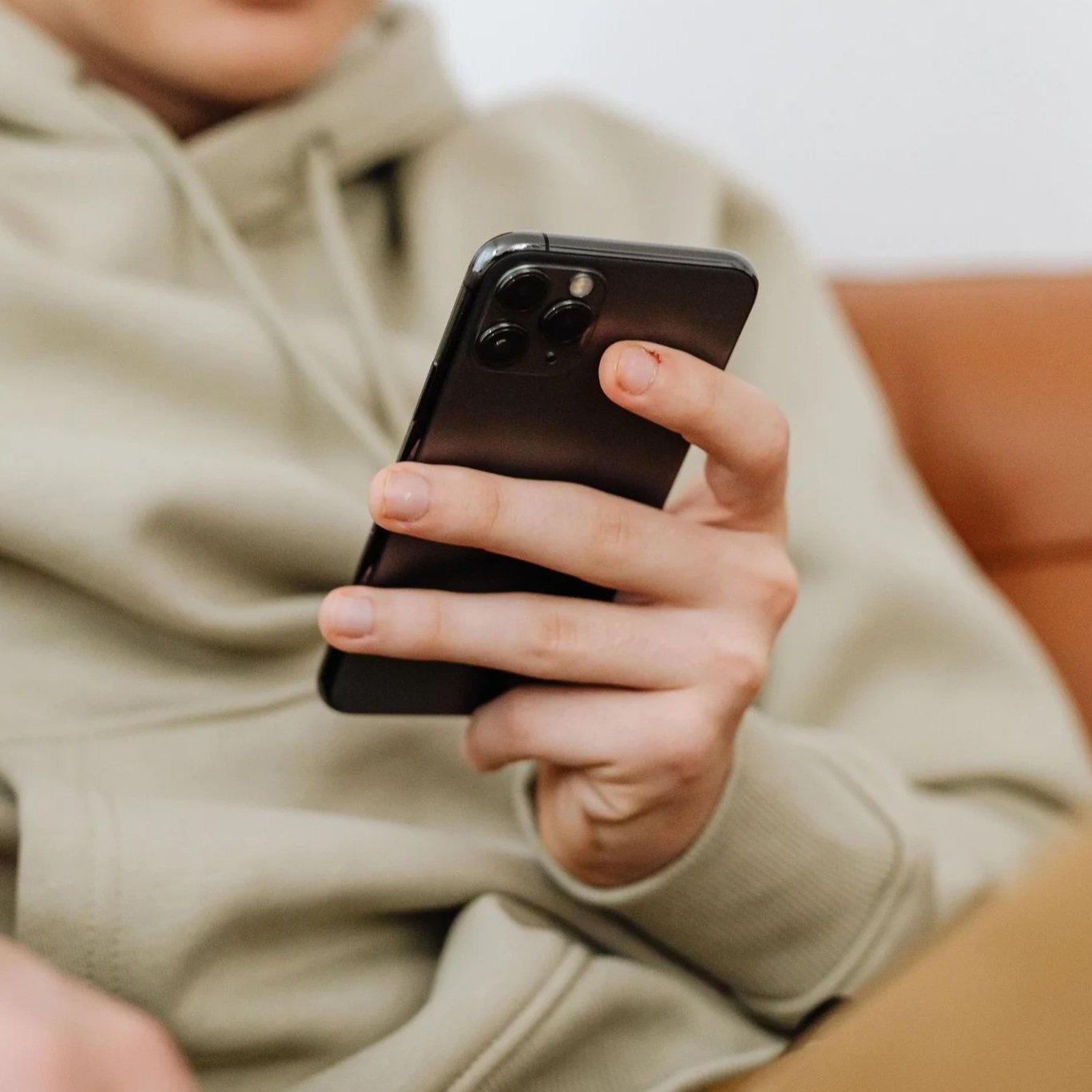 Person holding a black smartphone with a triple-camera setup, wearing a beige hoodie, sitting on a brown couch.