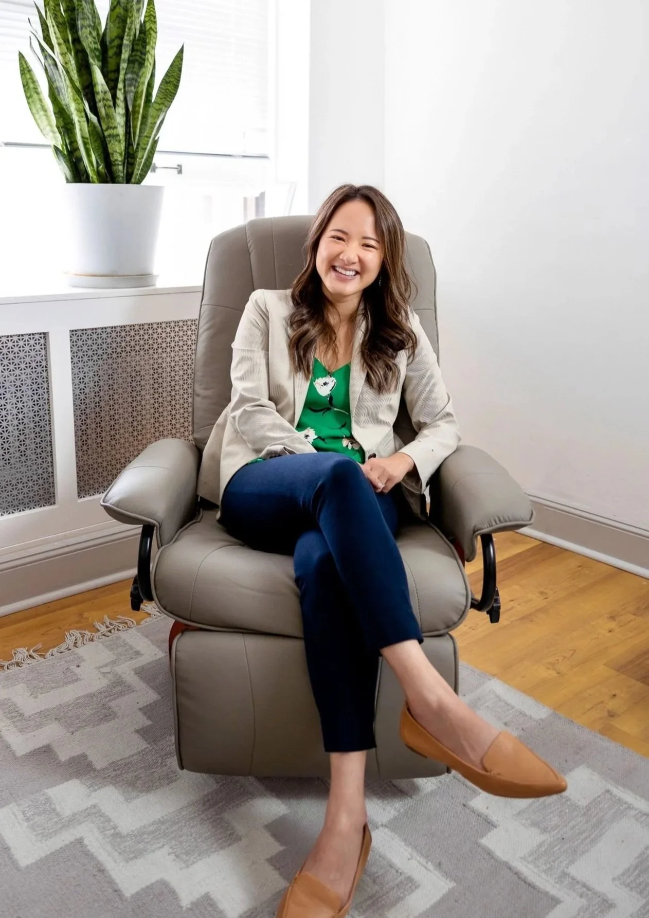 A woman Joanna Maung sitting in a beige armchair in a bright, modern room, smiling and laughing, with a large potted snake plant on the windowsill behind her, wearing a green blouse, beige blazer, dark jeans, and tan shoes.