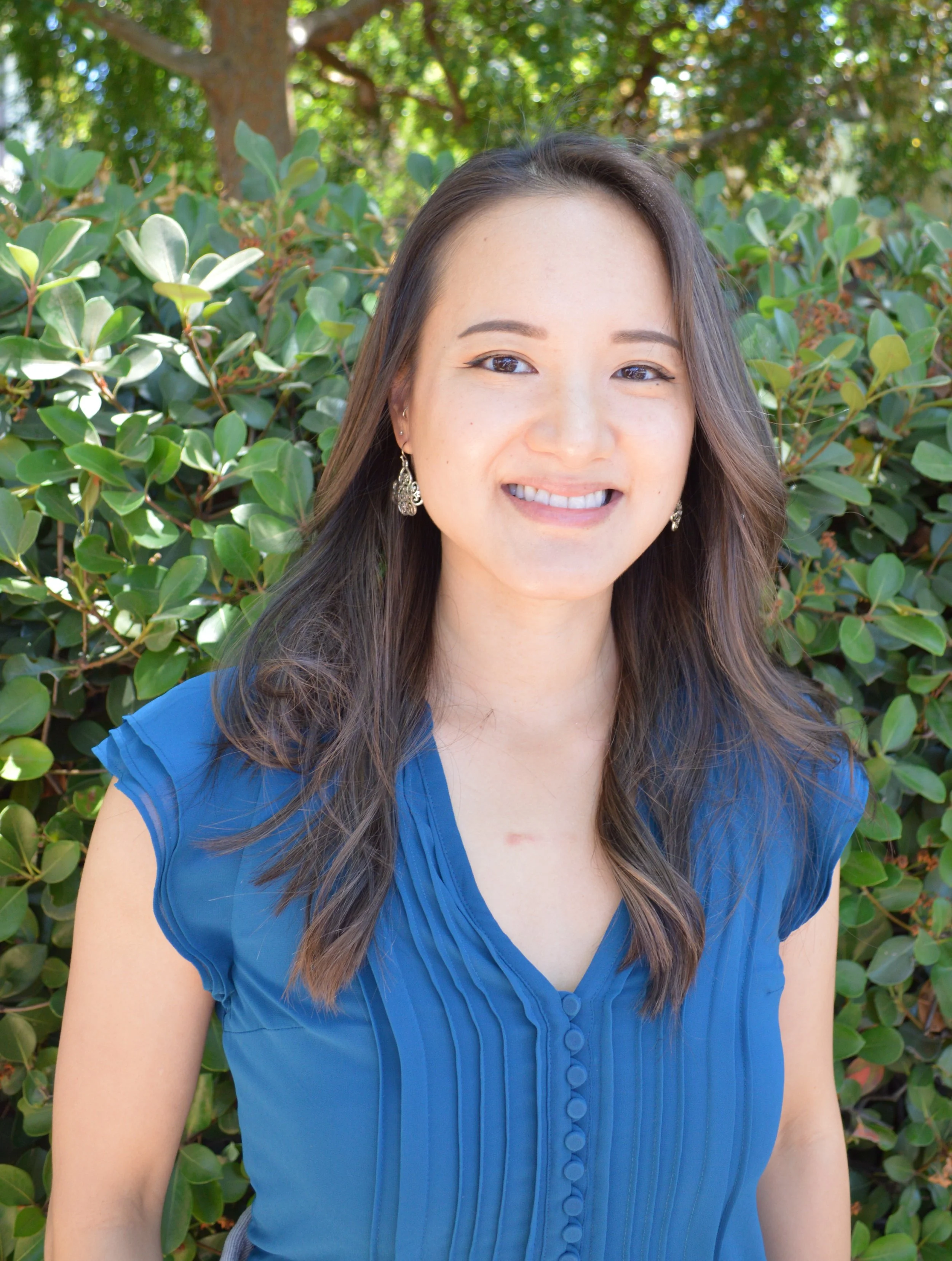 A woman with long dark hair wearing a blue blouse, smiling, standing outdoors in front of green foliage and trees.