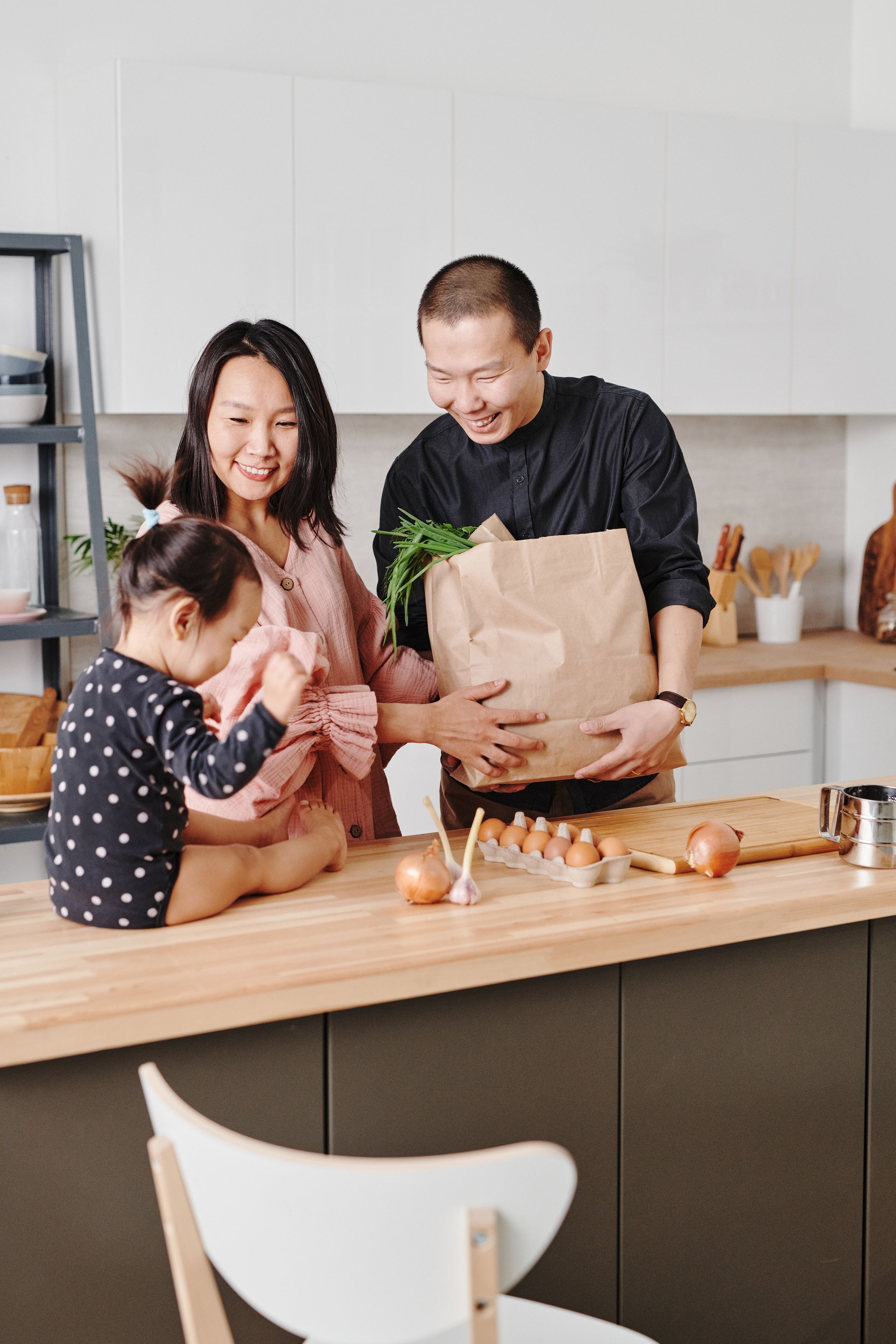 A family of three, including a mother, father, and daughter, preparing a meal together in a modern, white kitchen. The mother and father are smiling as they give a brown paper bag, filled with fresh greens, to each other. The daughter, with braided hair, is focused on a task on the wooden kitchen counter, which has eggs, garlic, onions, and cooking utensils on it.