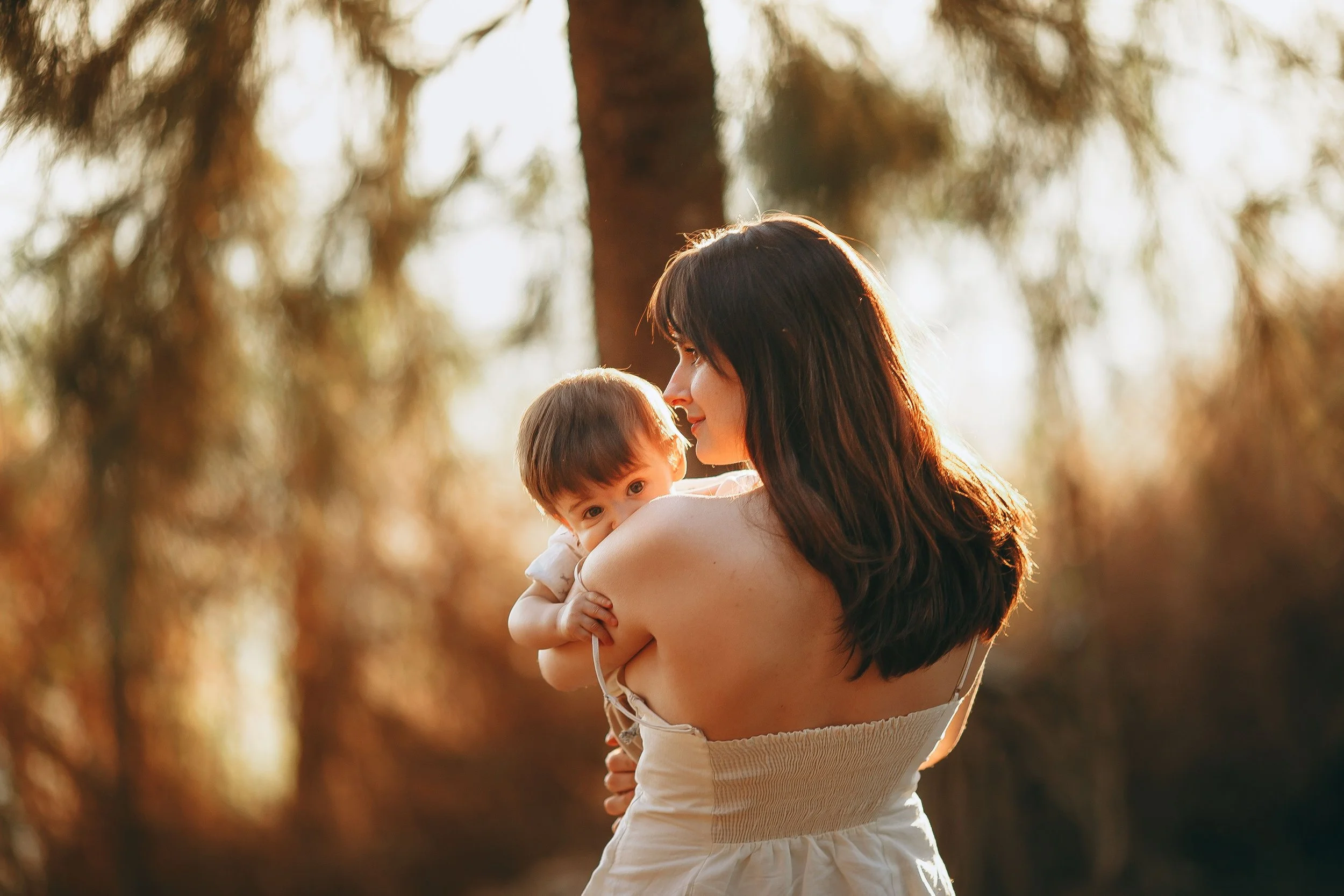 A woman with long dark hair smiling and holding a young child with brown hair outdoors in a wooded area during the golden hour, with warm sunlight filtering through trees.