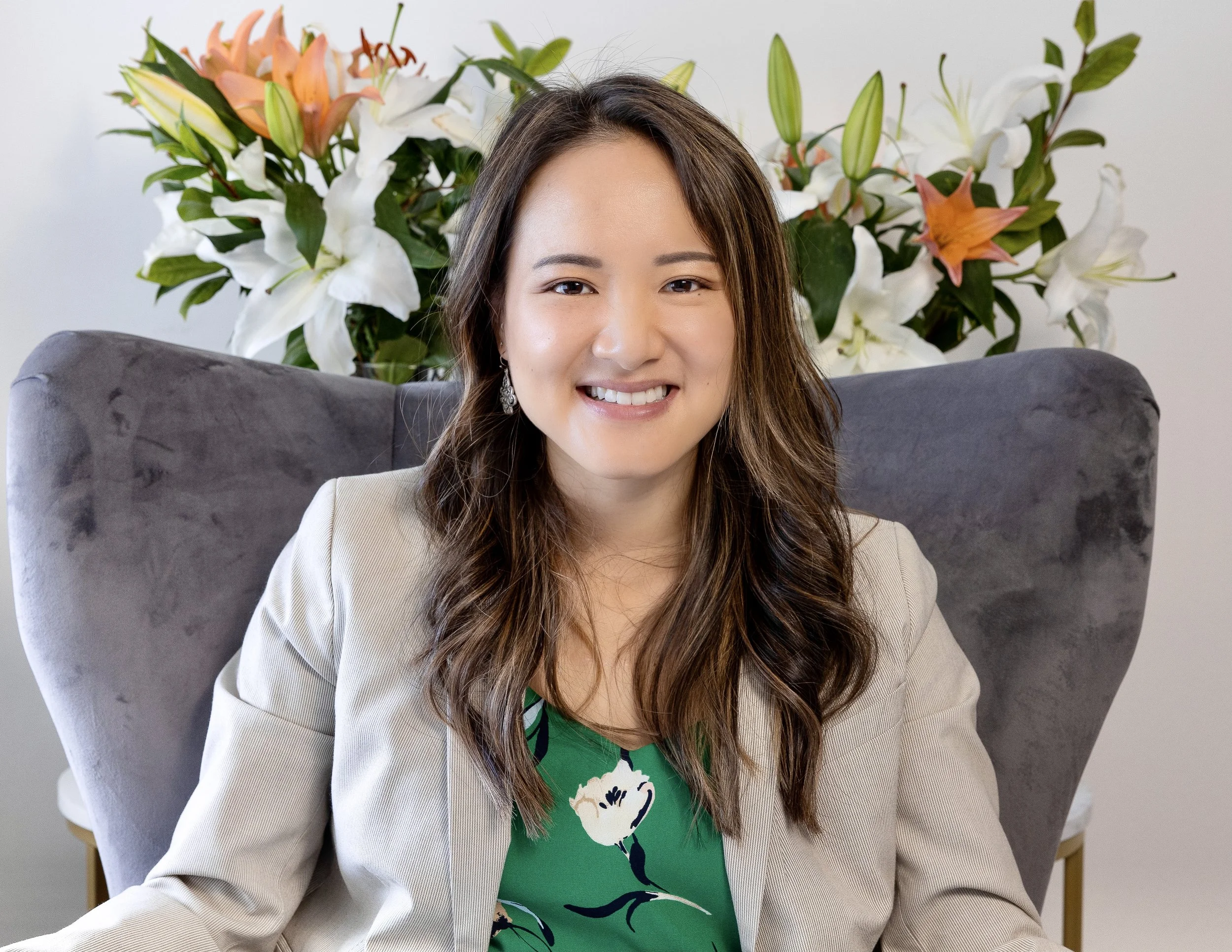 Portrait of Joanna Maung, a smiling woman with long brown hair, wearing a beige blazer over a green floral dress, seated on a gray upholstered chair with a large floral arrangement of white and orange lilies behind her.