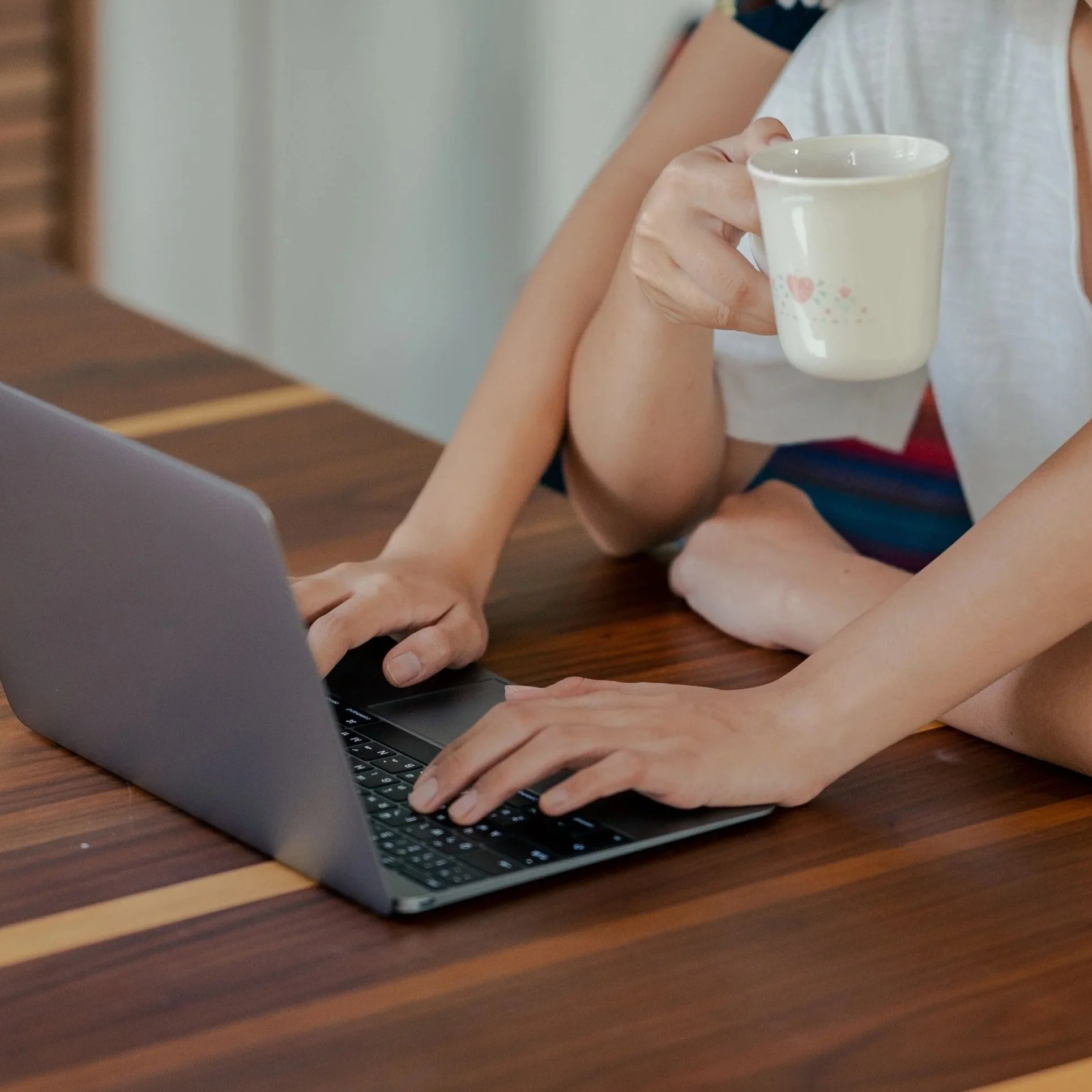 Person holding a mug while working on a laptop at a wooden table.