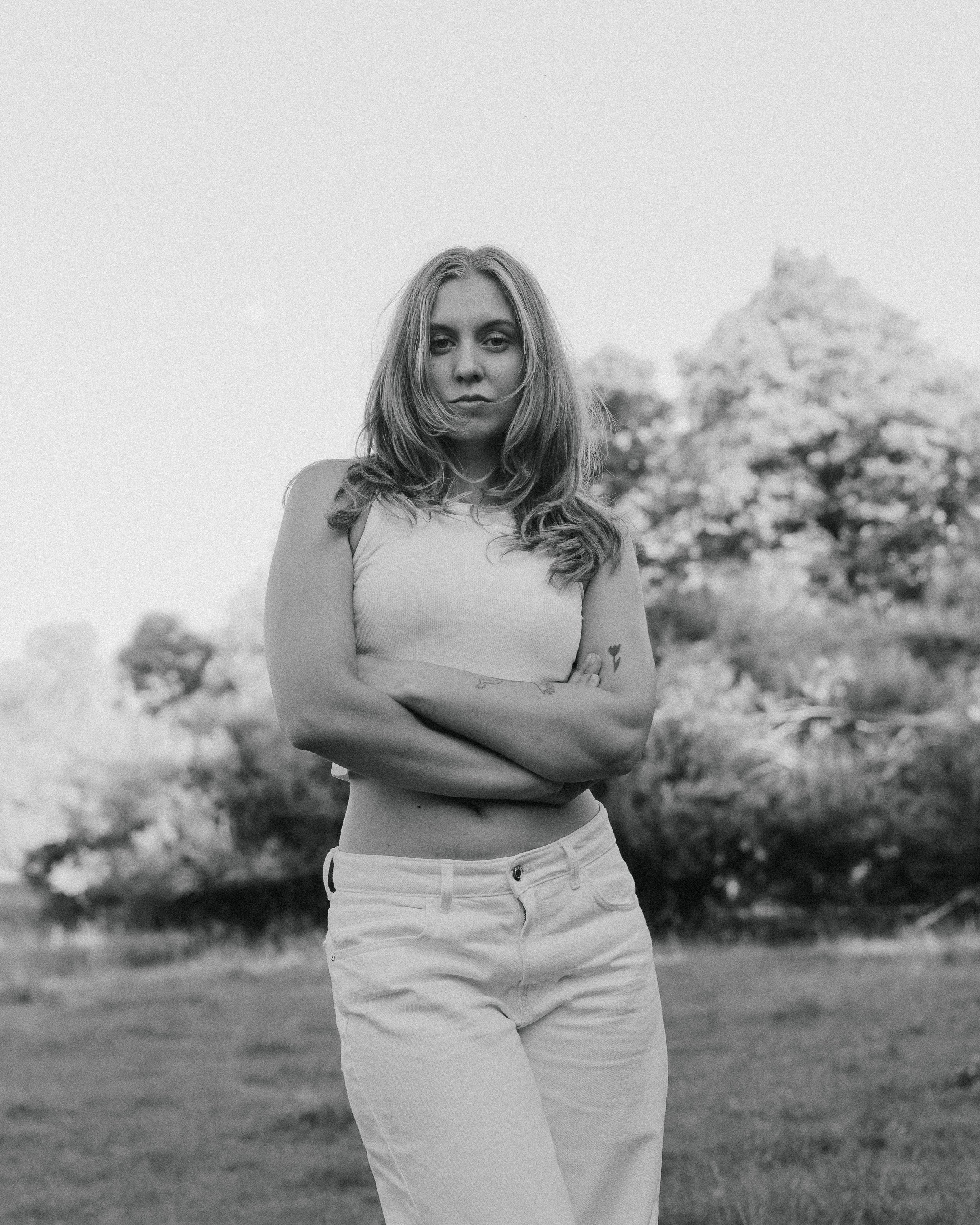 A woman with long wavy hair standing outdoors with a tree in the background, arms crossed, wearing a sleeveless top and white pants, in black and white.