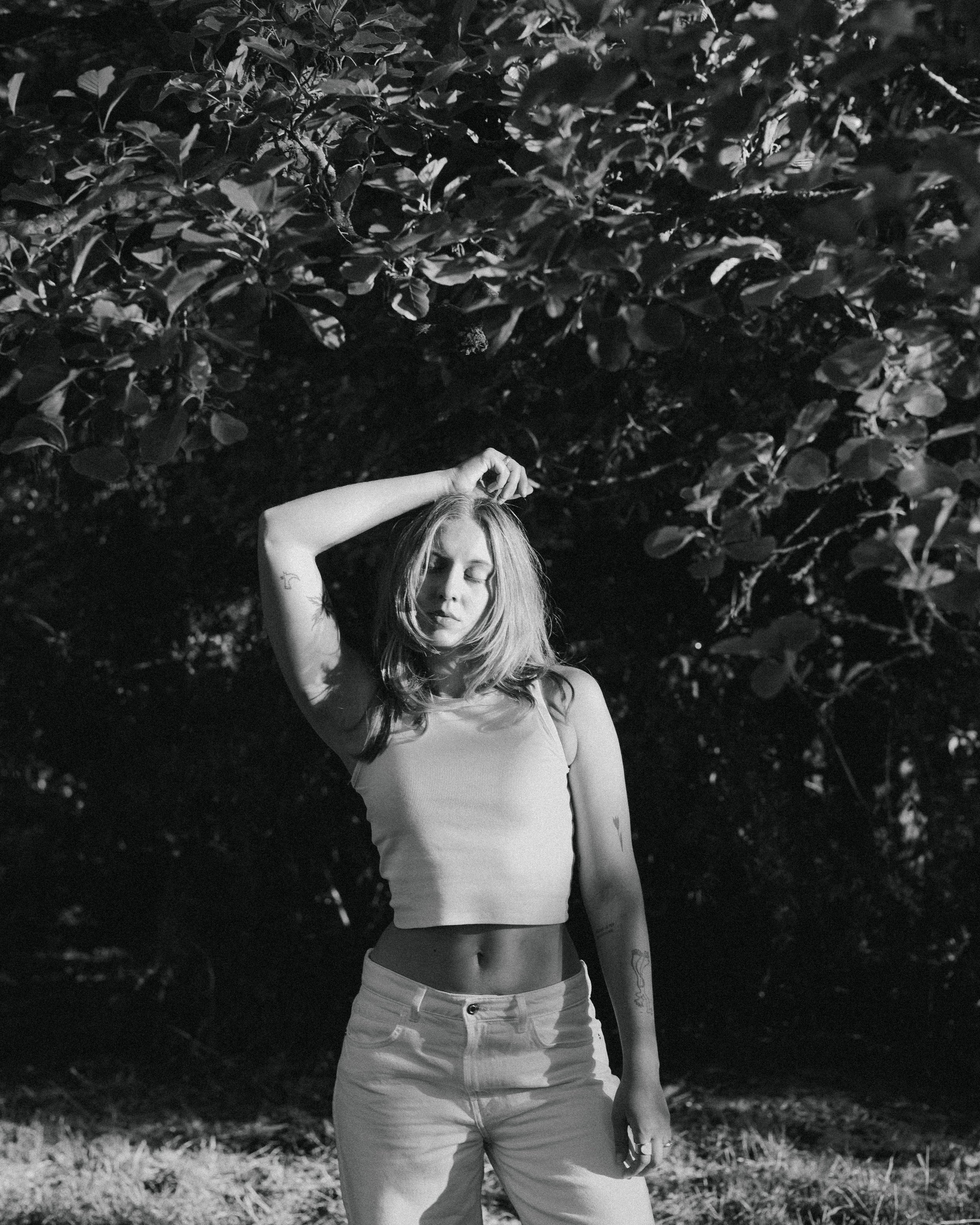 Black and white photograph of a woman standing outdoors at night, with her right hand raised to her head and her eyes closed. She is wearing a sleeveless top and jeans, and has visible tattoos on her left arm. Dense foliage is in the background.