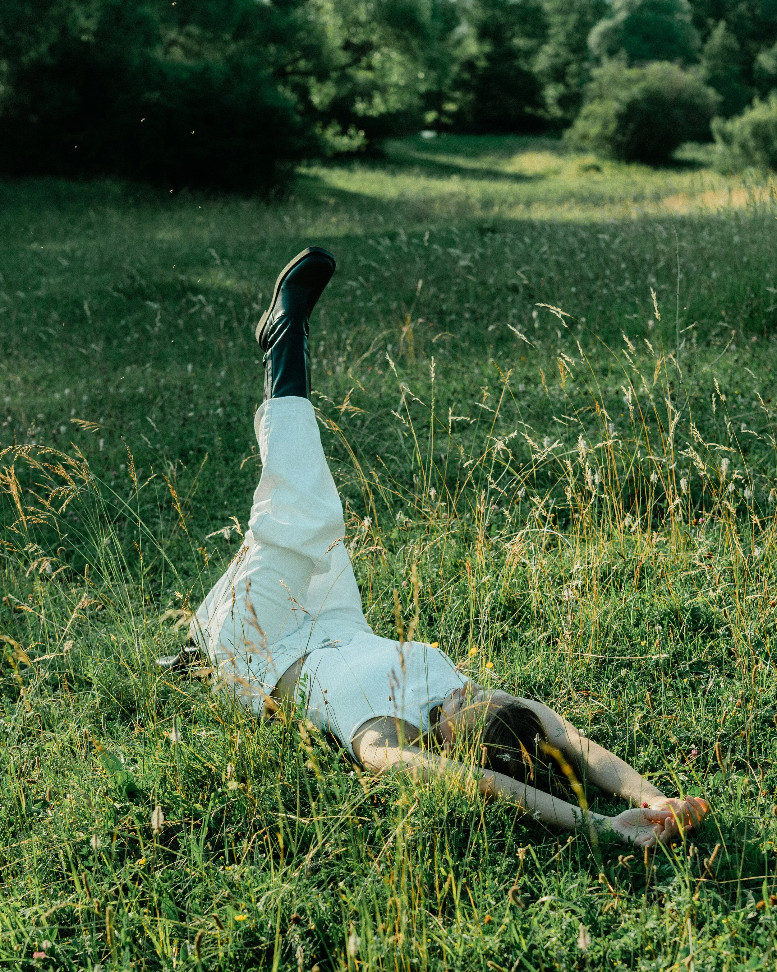 Person lying on grass in a field with legs raised, surrounded by tall grass and trees in the background.