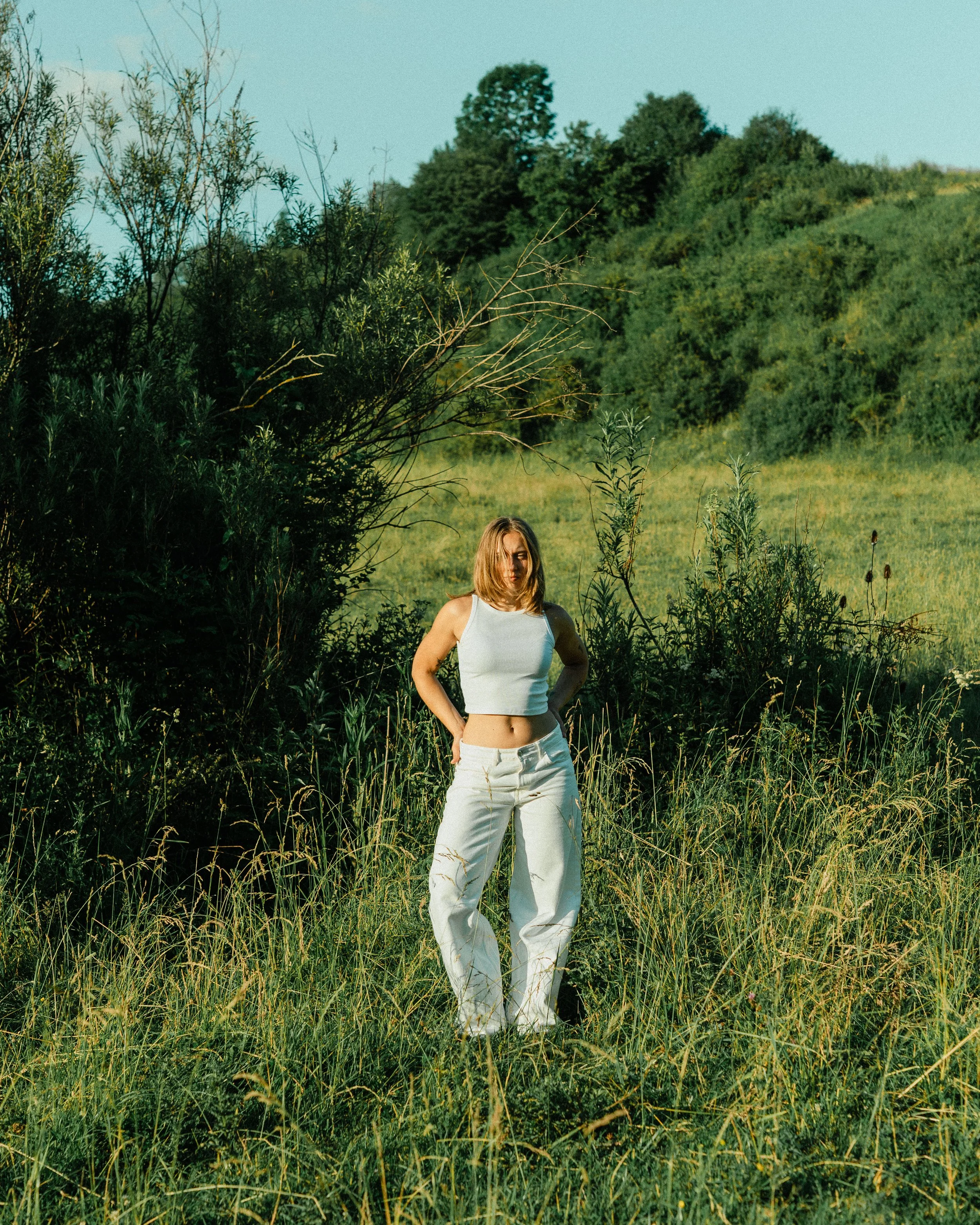 A woman with blonde hair wearing a white tank top and white pants standing in a grassy field with trees and hills in the background.