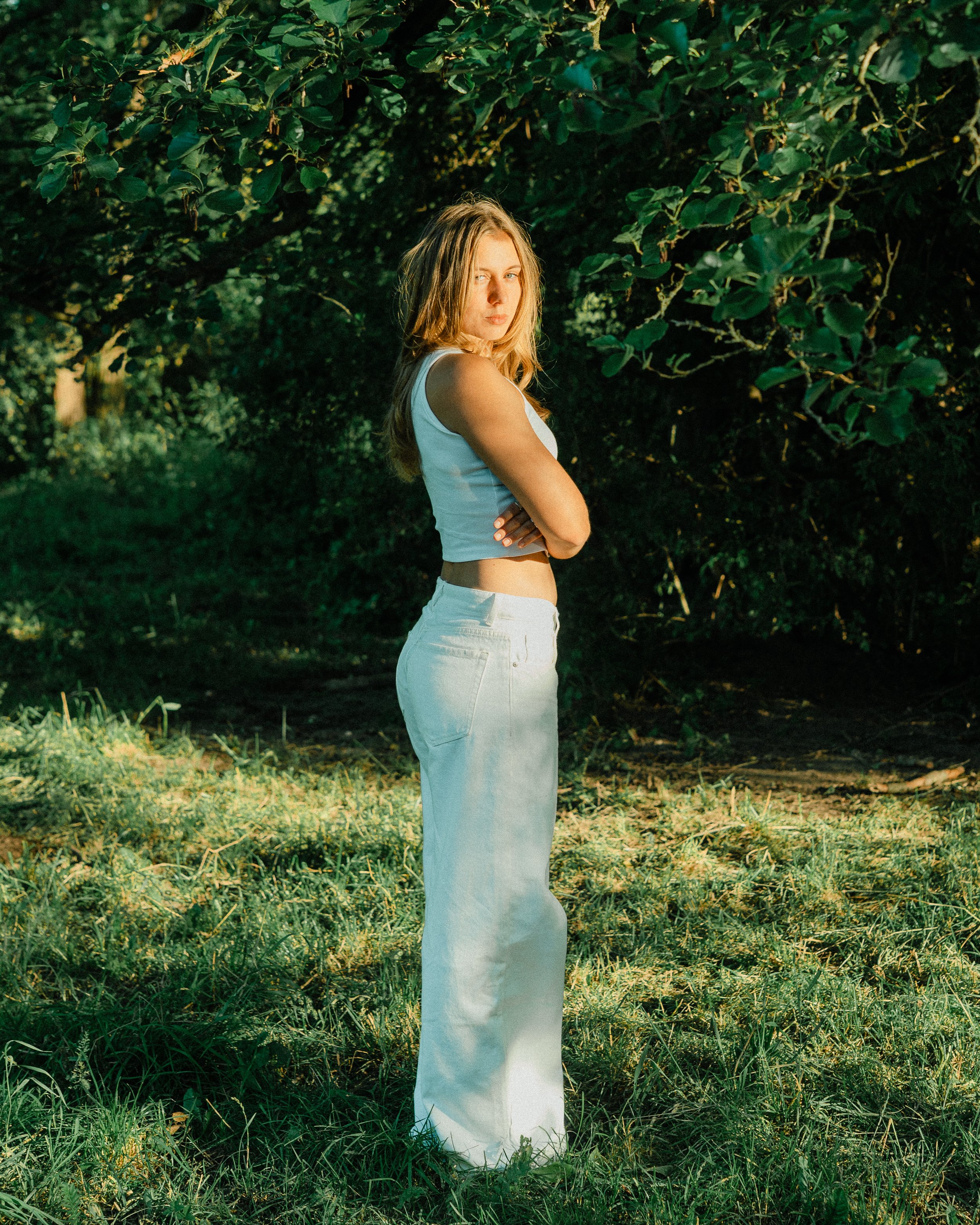 A young woman with blonde hair wearing a white tank top and white jeans standing with arms crossed in front of green leafy trees and grass.