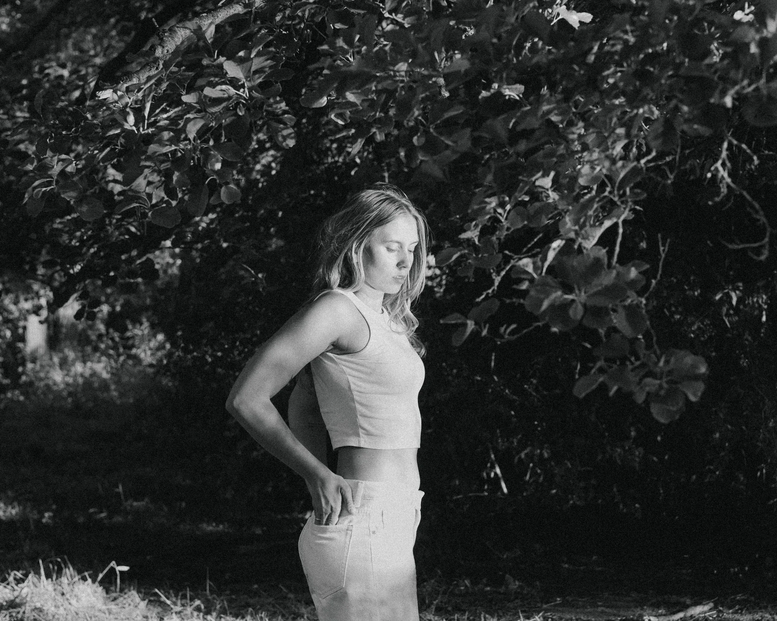 Black and white photo of a young woman with long hair standing outdoors, with trees and foliage in the background. She is wearing a sleeveless top and jeans, looking down with her eyes closed.
