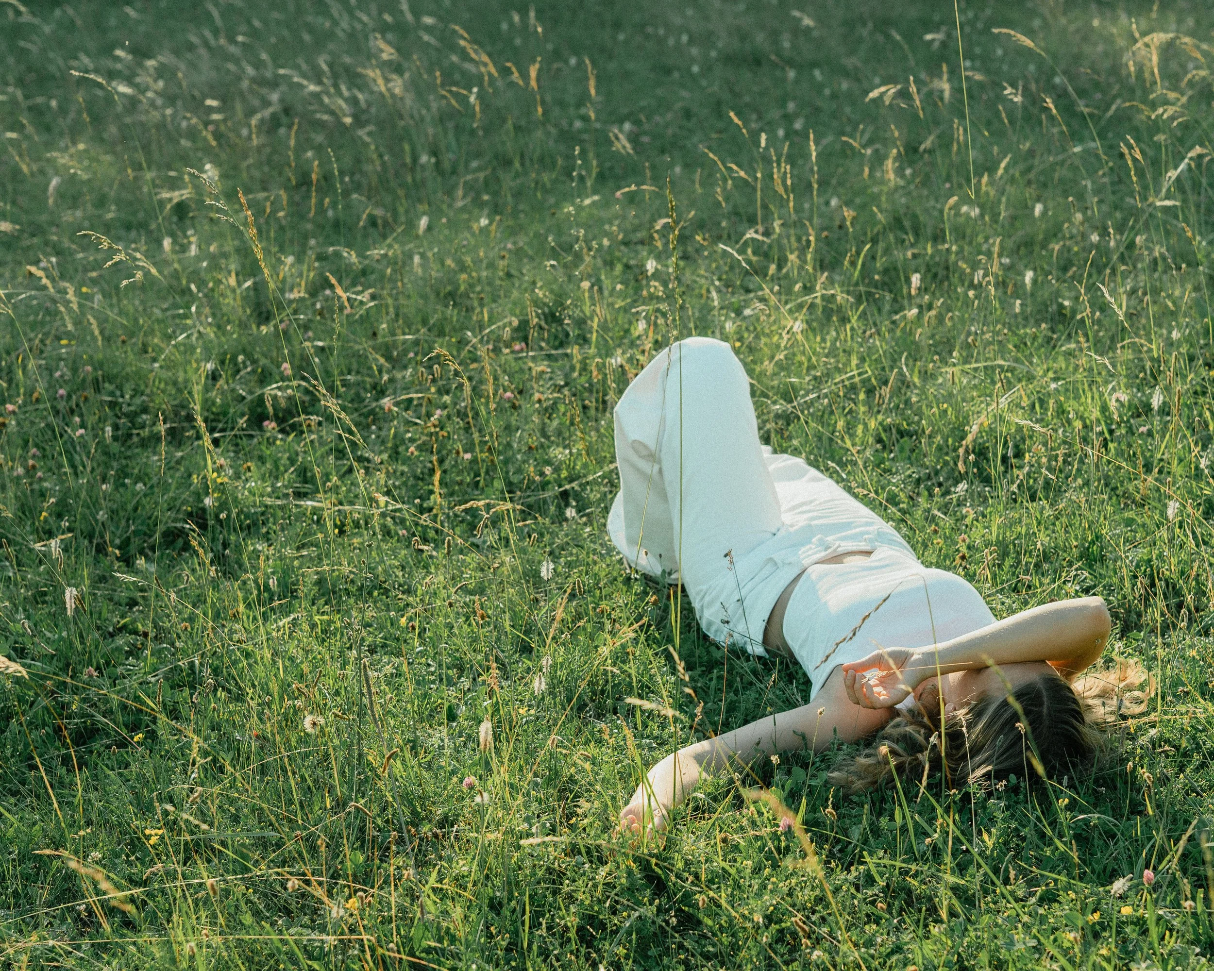 Person lying on grass in a field, wearing white clothing, with their head turned to the side and one arm extended.