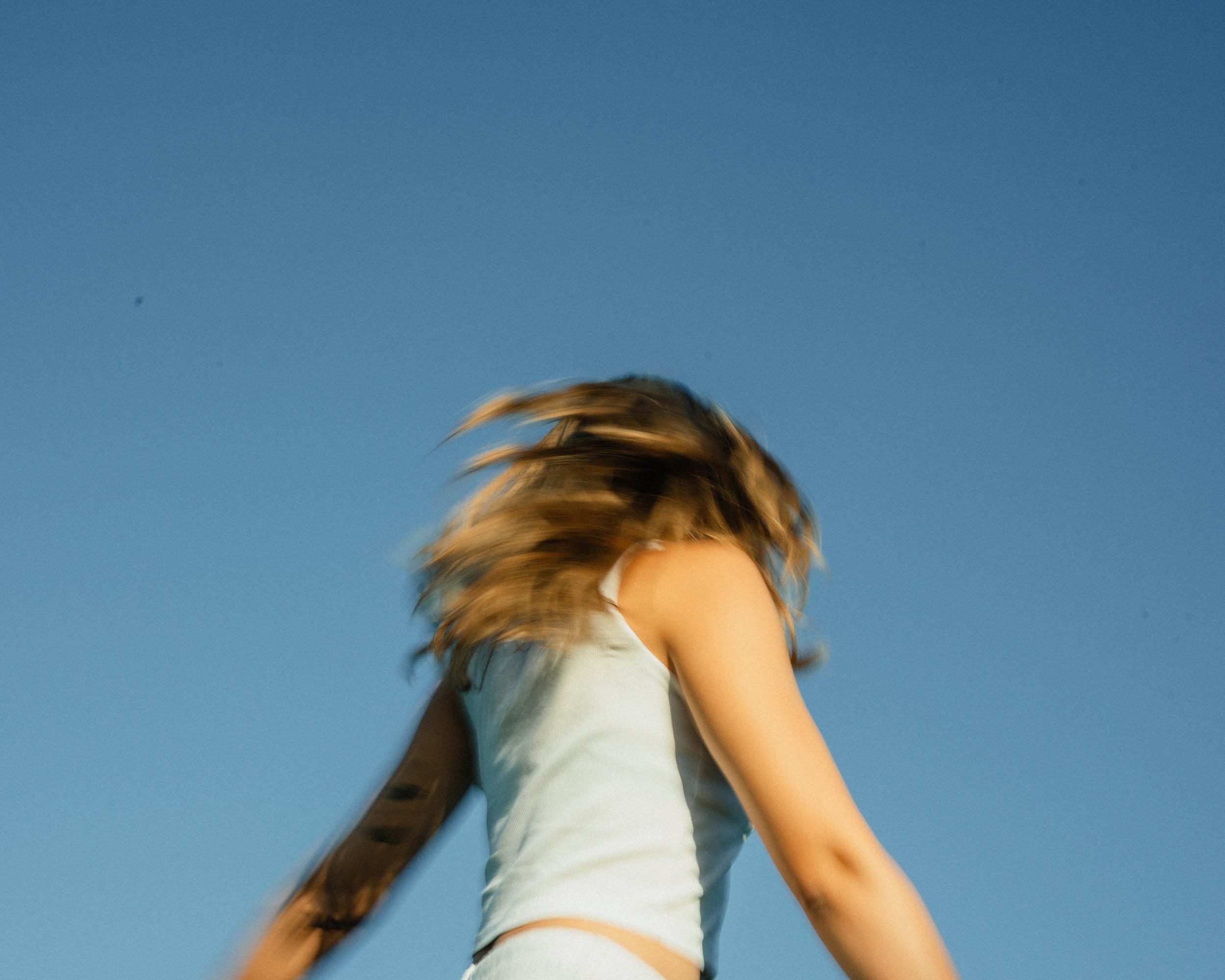 Part of a person with brown hair wearing a white t-shirt, standing outdoors against a clear blue sky.