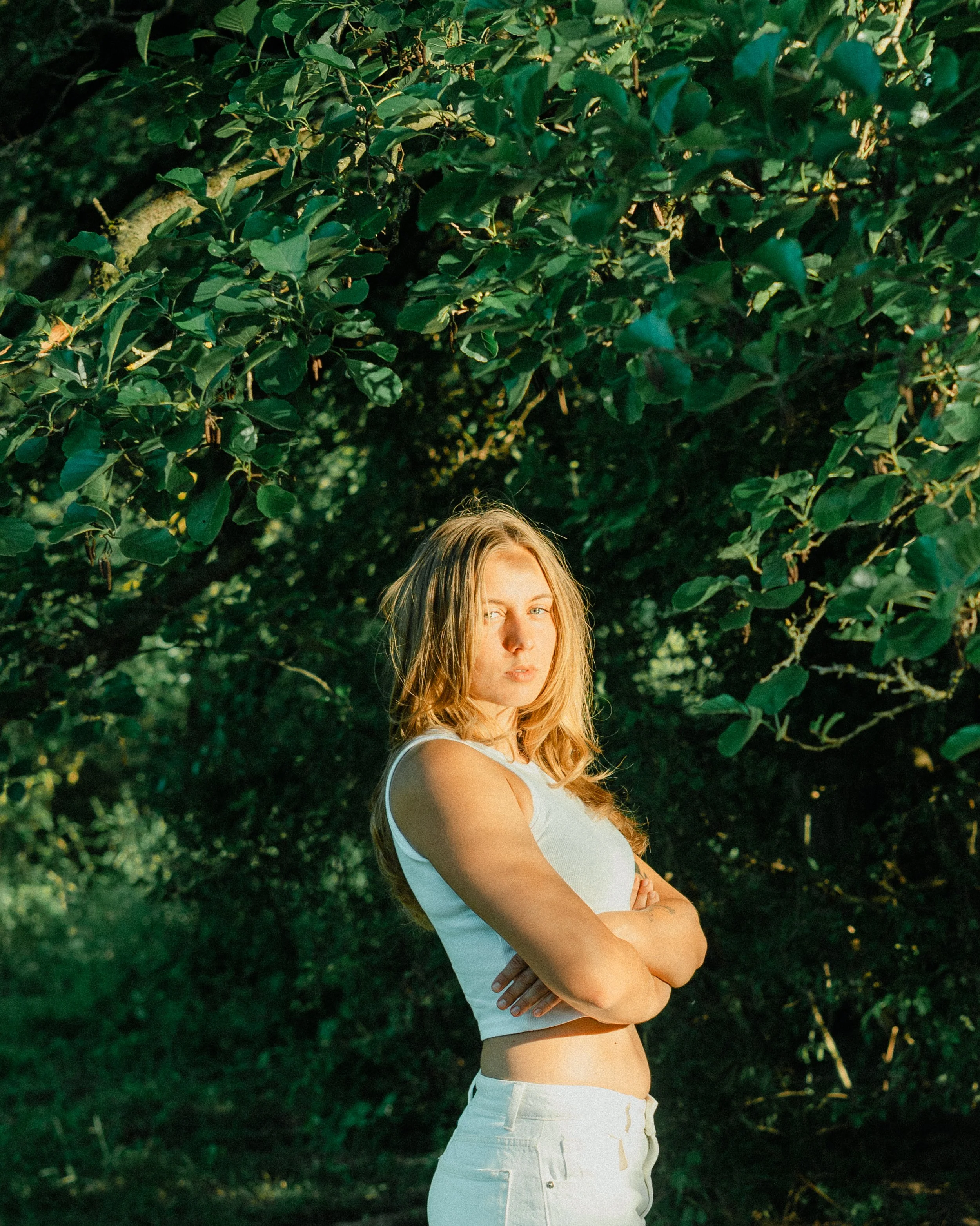 A woman with long blonde hair standing outdoors against a background of dense green foliage, wearing a white sleeveless top and white shorts, with her arms crossed and looking slightly to the side.