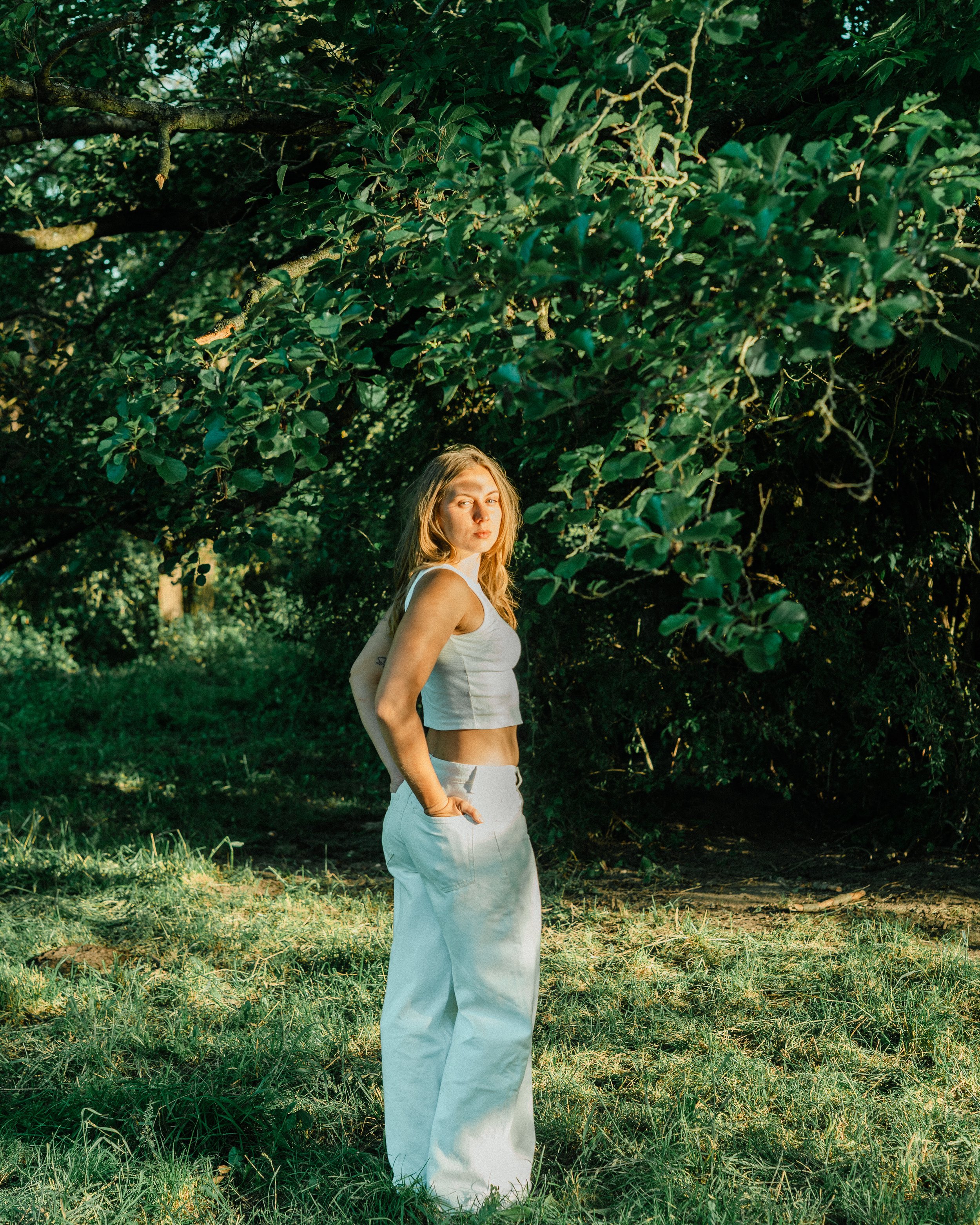 A woman with long blonde hair and light skin wearing a white sleeveless top and white pants stands outdoors in a green, leafy area, looking at the camera with a serious expression.