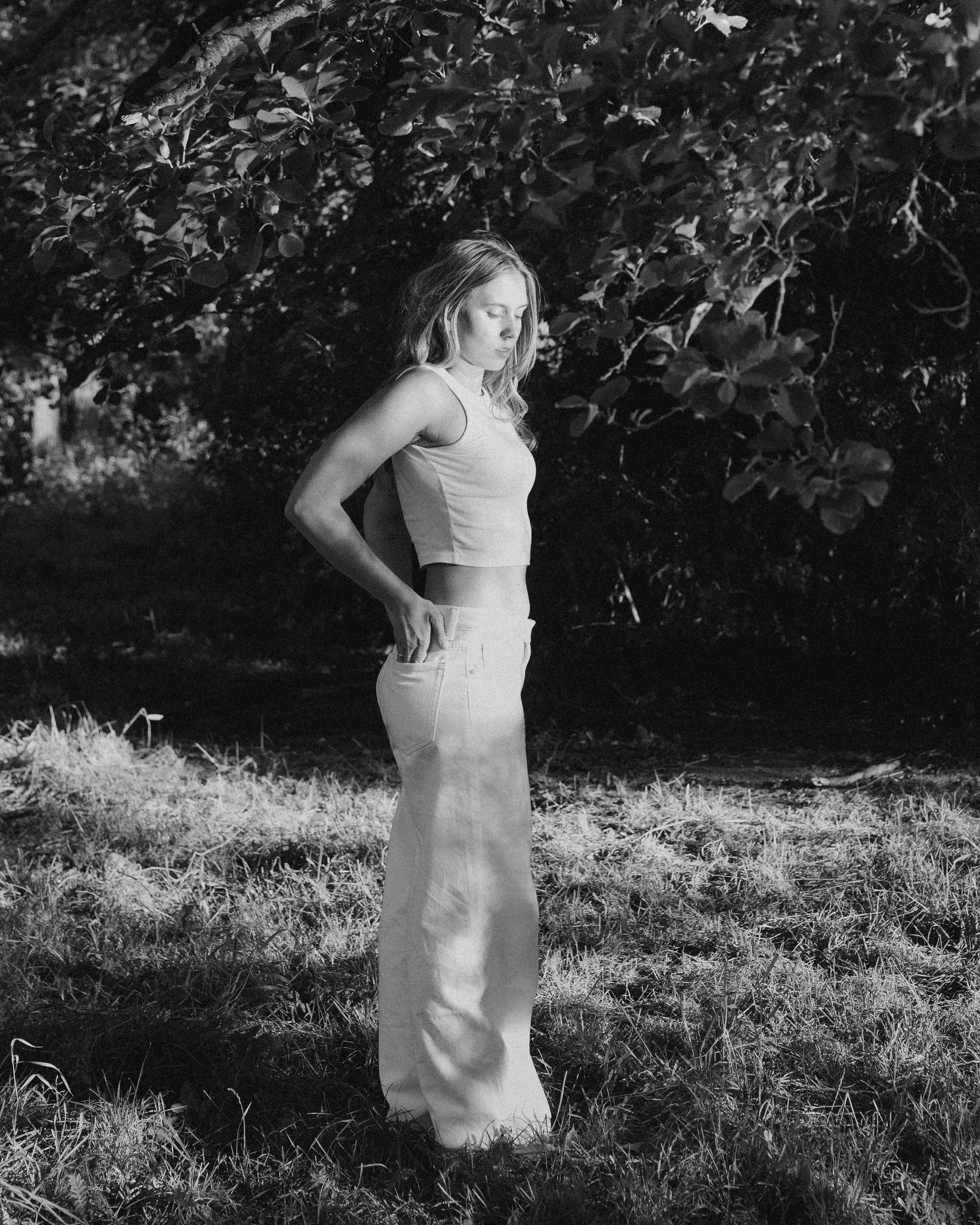 A black-and-white photograph of a young woman standing in a grassy outdoor setting with dense foliage and trees in the background. She is wearing a sleeveless top and high-waisted pants, with her eyes closed and one hand resting in her pocket.