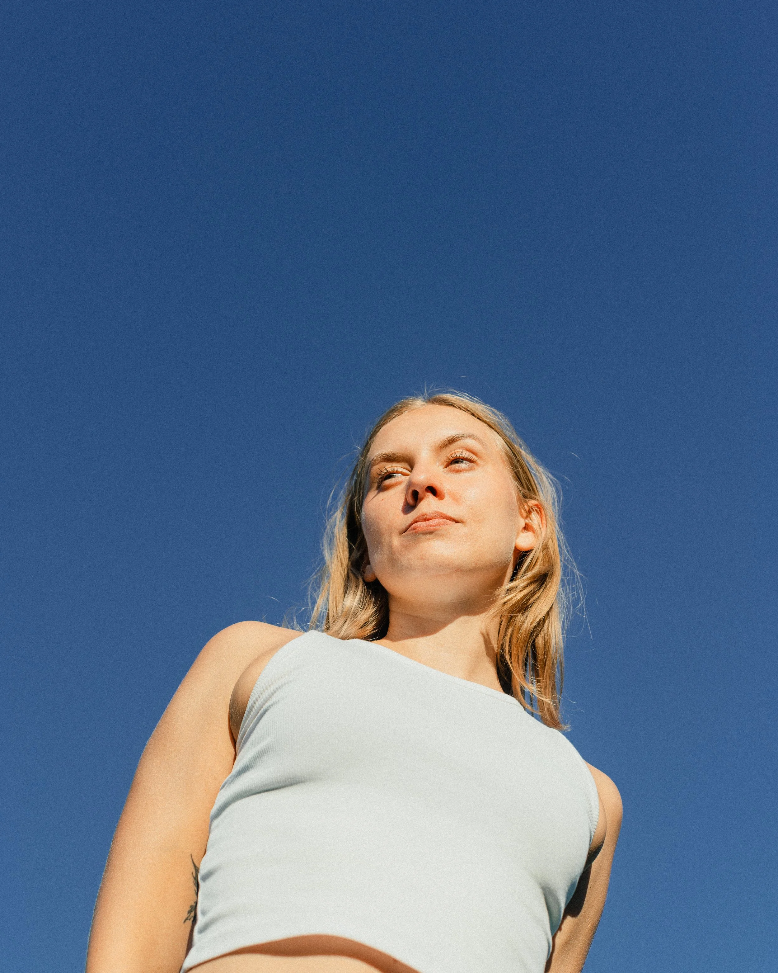 A young woman with blonde hair looking slightly upward, wearing a white top, against a clear blue sky.