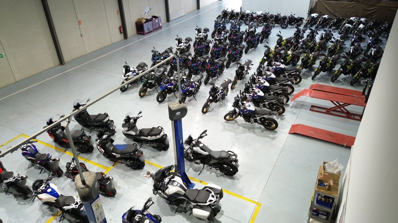 Indoor parking area filled with numerous motorcycles and scooters arranged in rows, with some red platform lifts and a few small storage shelves in the background.