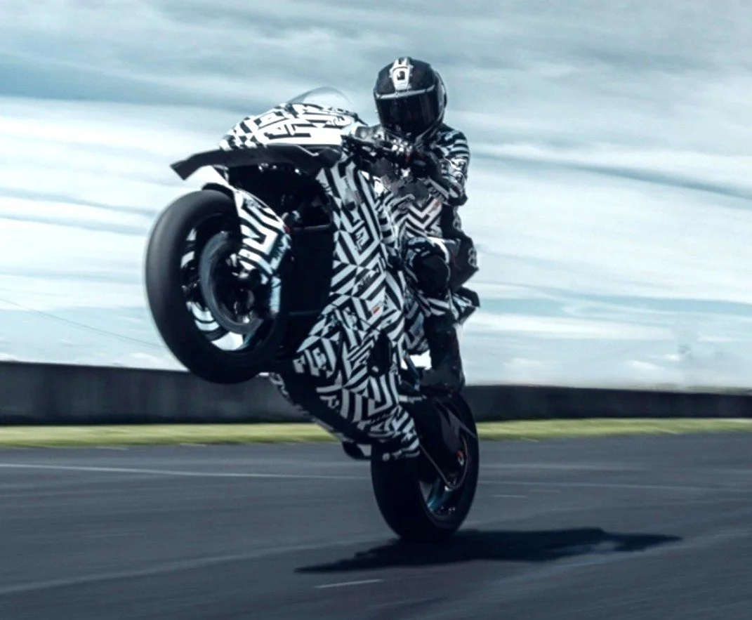 A person wearing a helmet and camouflage-patterned racing suit performing a wheelie on a motorcycle on a race track.